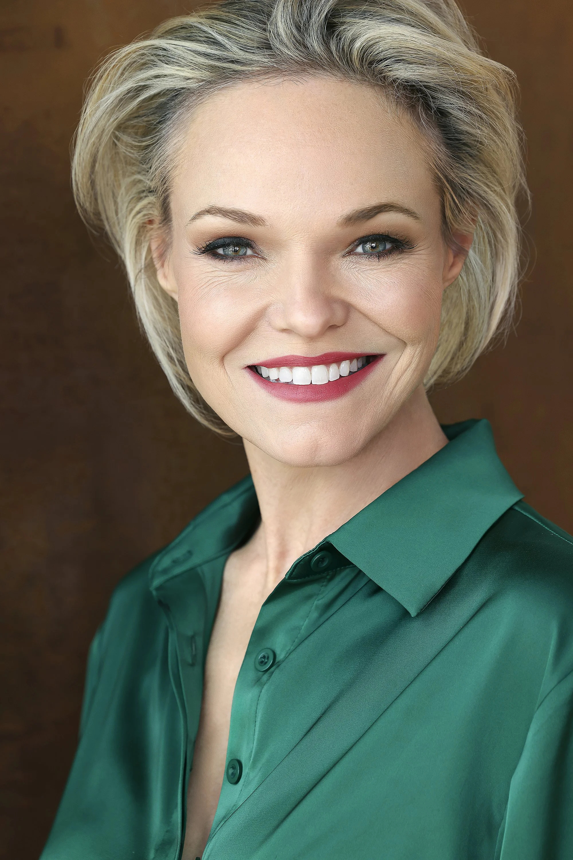 A smiling middle-aged woman with short blonde hair, blue eyes, wearing a green button-up shirt, against a brown background.