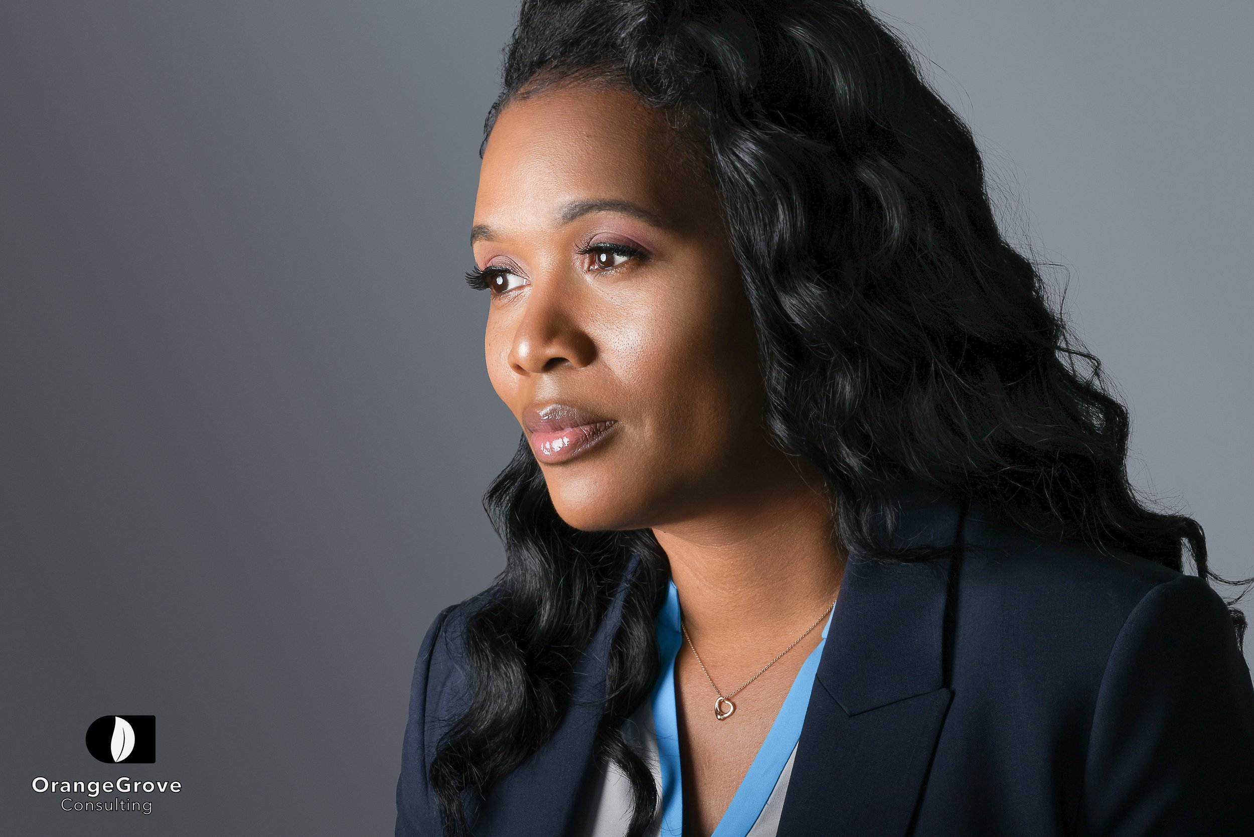 A professional woman with long, wavy black hair wearing a dark blazer and a necklace, looking to the side against a plain gray background. Logo in lower left corner reads 'OrangeGrove Consulting'.