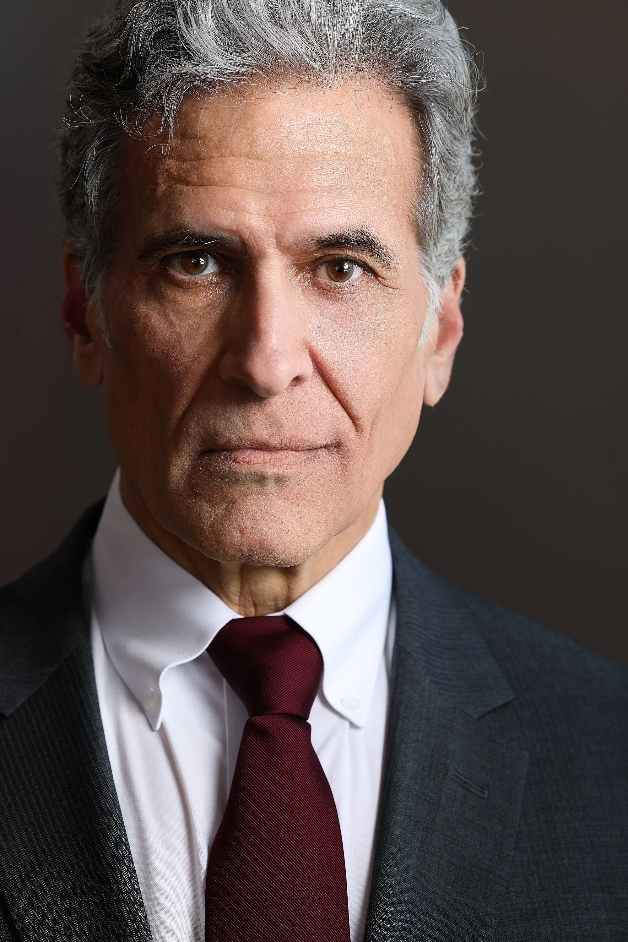 Close-up portrait of a mature man with gray hair and brown eyes, wearing a dark suit, white shirt, and maroon tie, looking directly at the camera against a dark background.