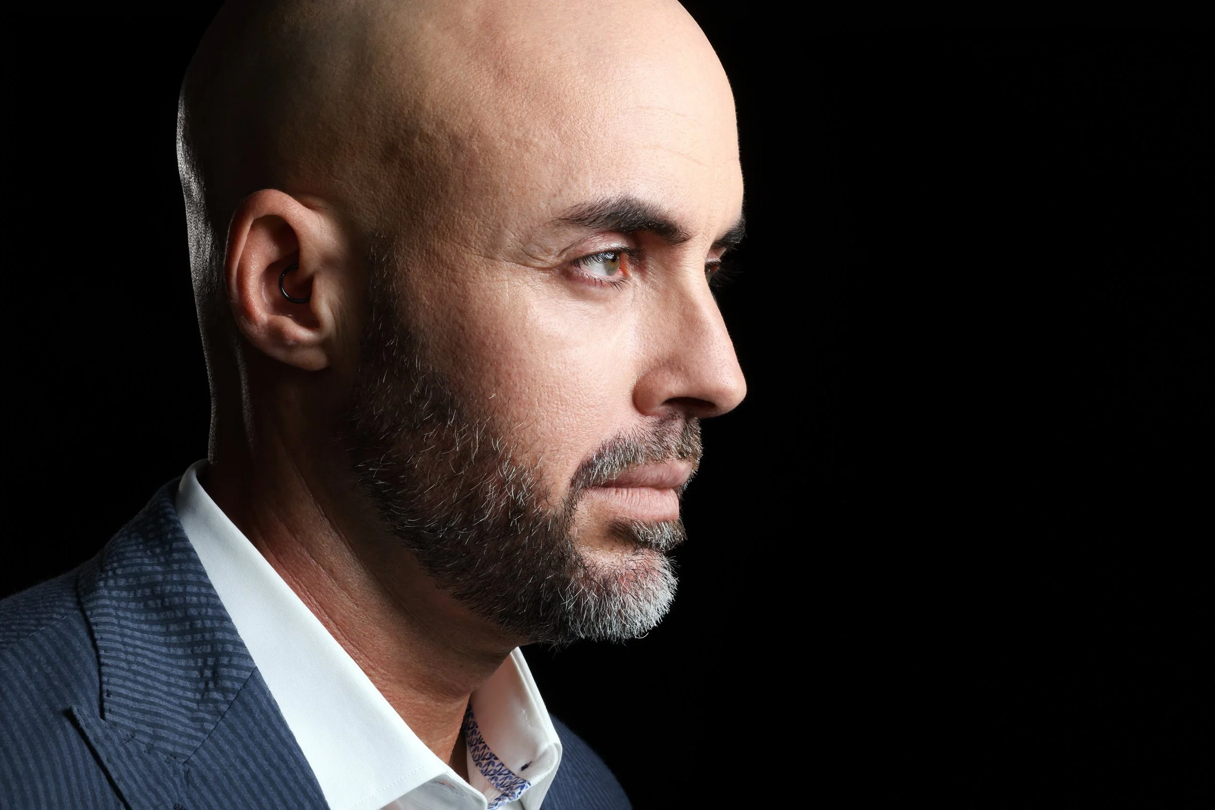 Close-up profile of a bald man with a beard, wearing a dark blue suit and white shirt, against a black background.