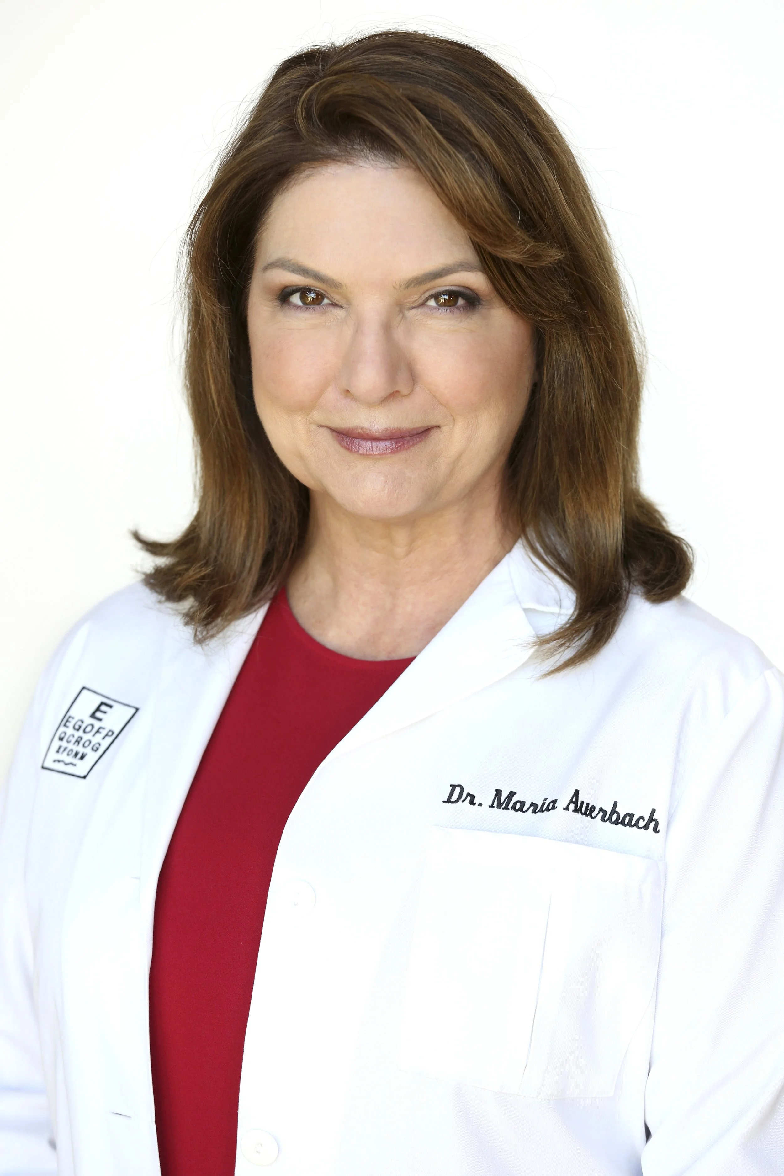 A woman with shoulder-length brown hair wearing a white doctor's coat with her name, Dr. Maria Auerbach, embroidered on it, and a red shirt underneath, standing against a plain white background.