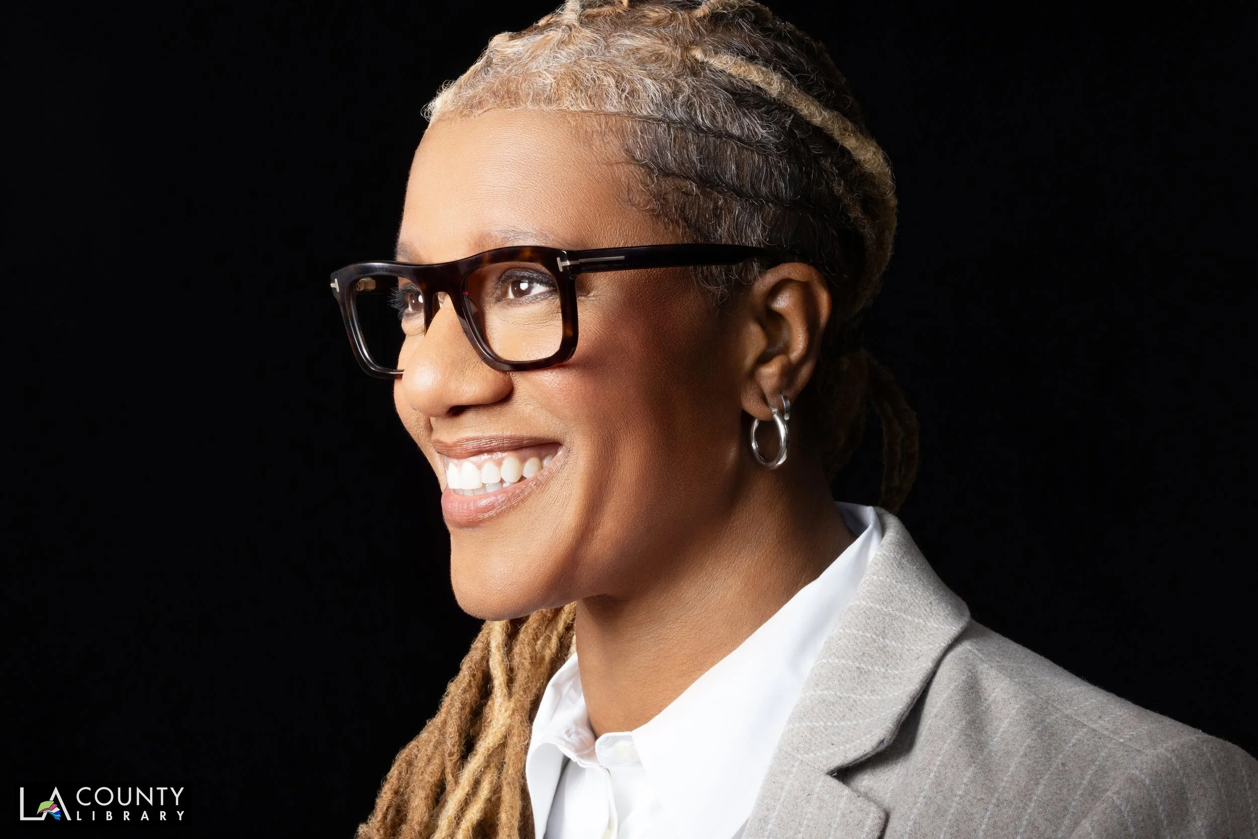 A smiling woman with light brown skin, wearing black glasses, hoop earrings, a gray pinstripe blazer, and a white shirt, against a black background.