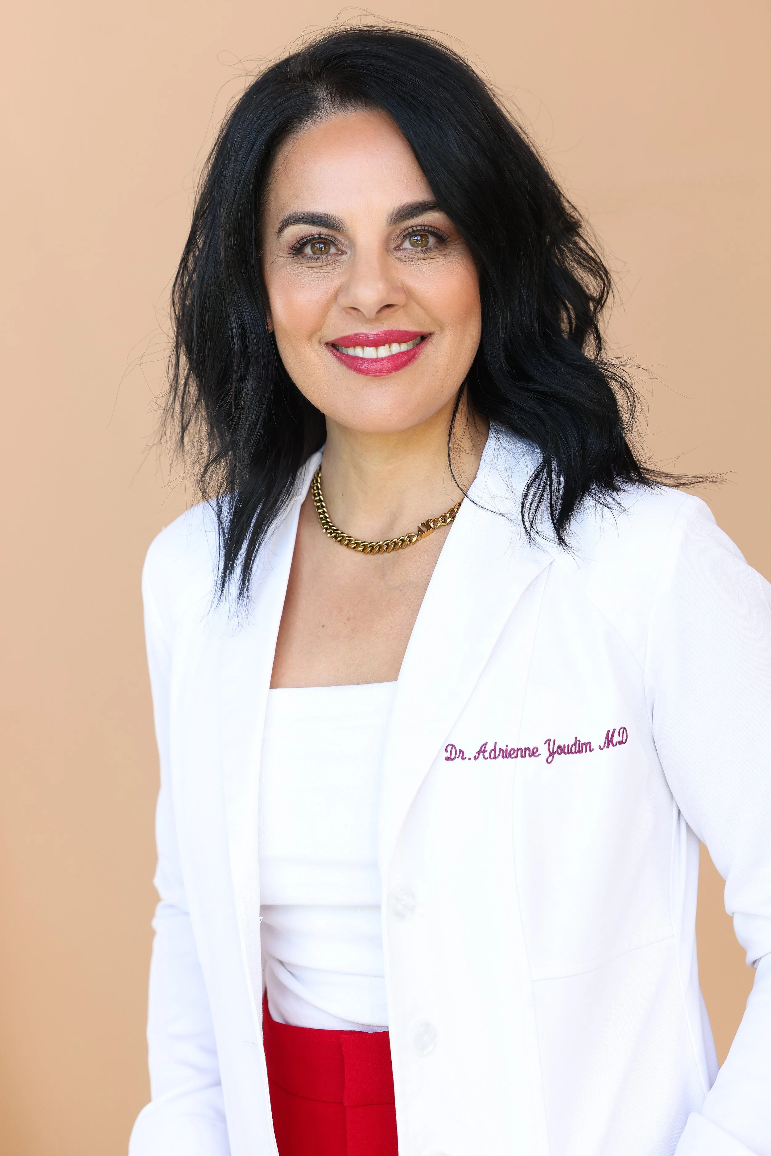 A woman with shoulder-length black hair and light makeup, wearing a white medical coat with embroidered text, a gold necklace, and red clothing underneath, standing against a peach-colored background, smiling at the camera.