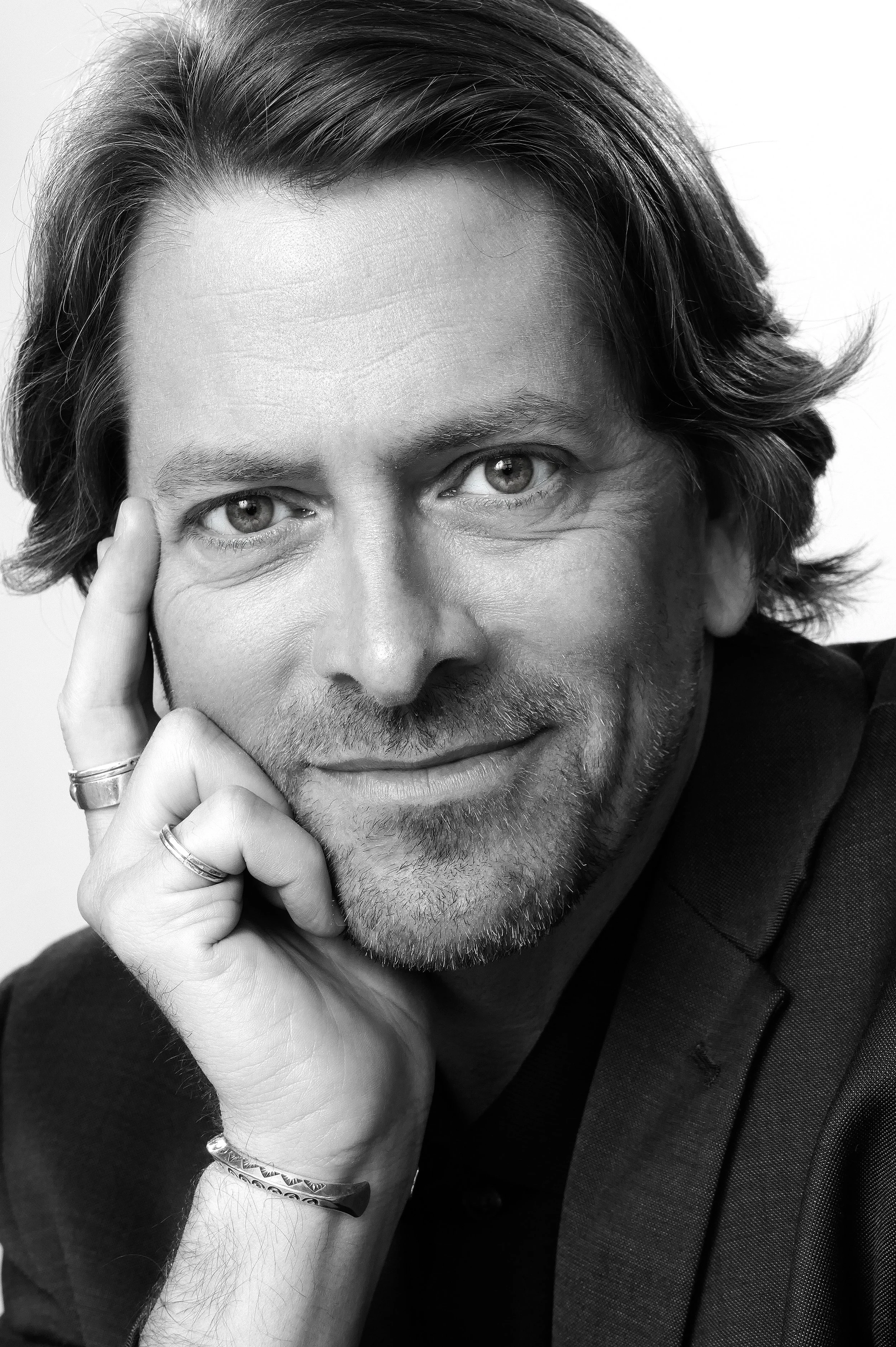 A black and white portrait of a man with medium-length hair, light-colored eyes, and a slight beard. He is smiling slightly, resting his face on his left hand, with his left index finger touching his temple. He is wearing rings on his fingers, a brac