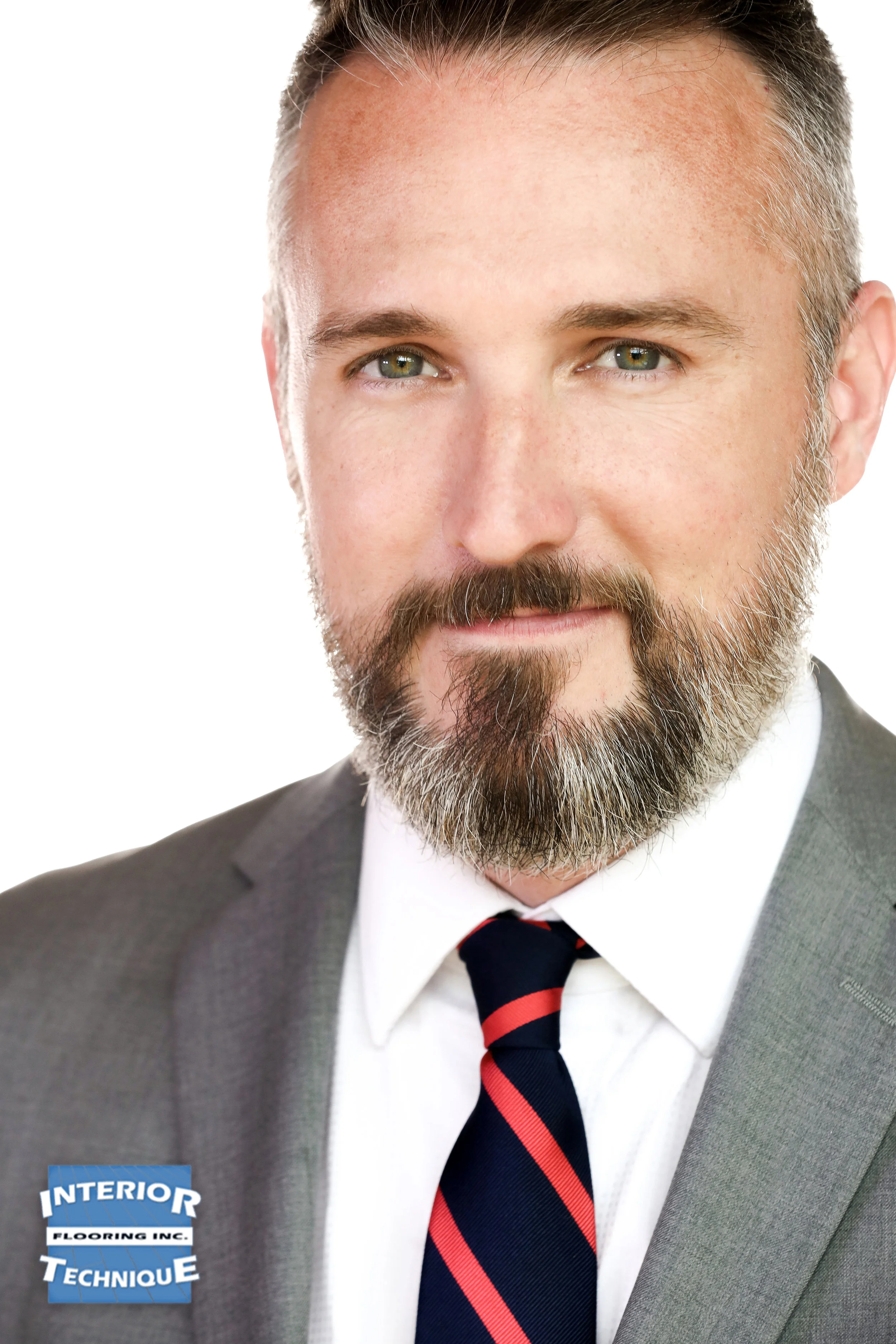 Portrait of a man with a beard and gray hair, wearing a gray suit, white shirt, and a navy tie with red stripes, against a white background.