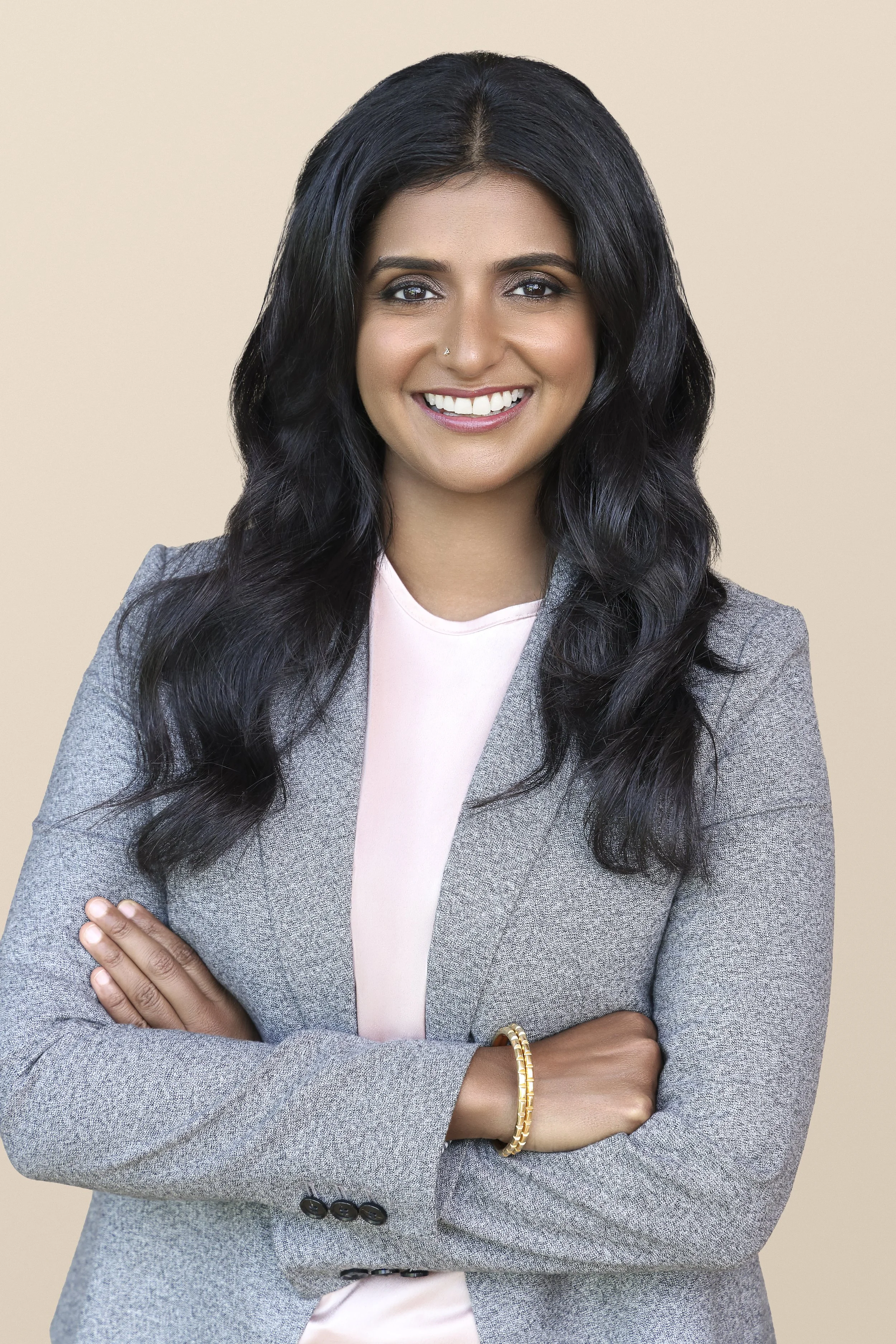 A woman with long, wavy black hair and a bright smile, wearing a gray blazer and a light pink shirt, standing with arms crossed against a neutral beige background.