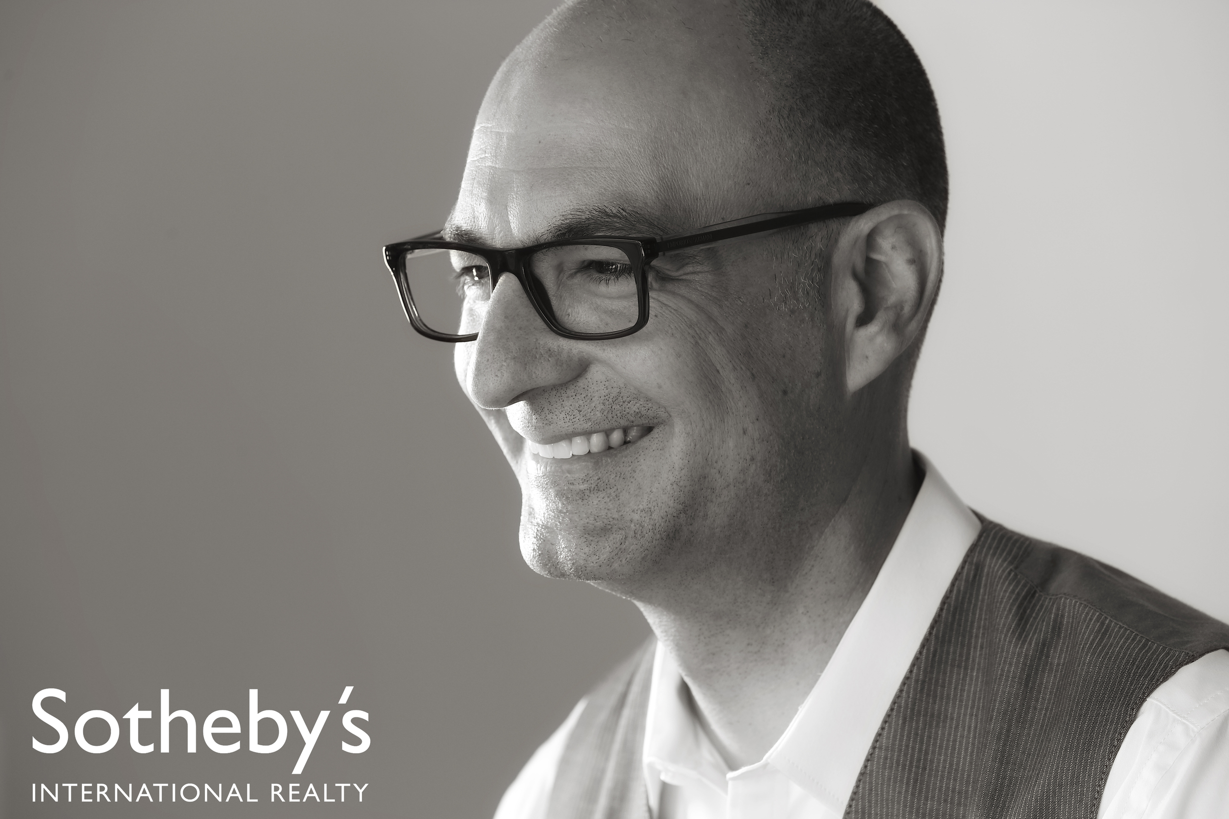 A smiling man with glasses, wearing a dress shirt and vest, in a black and white photo. The text 'Sotheby's International Realty' is in the bottom left corner.