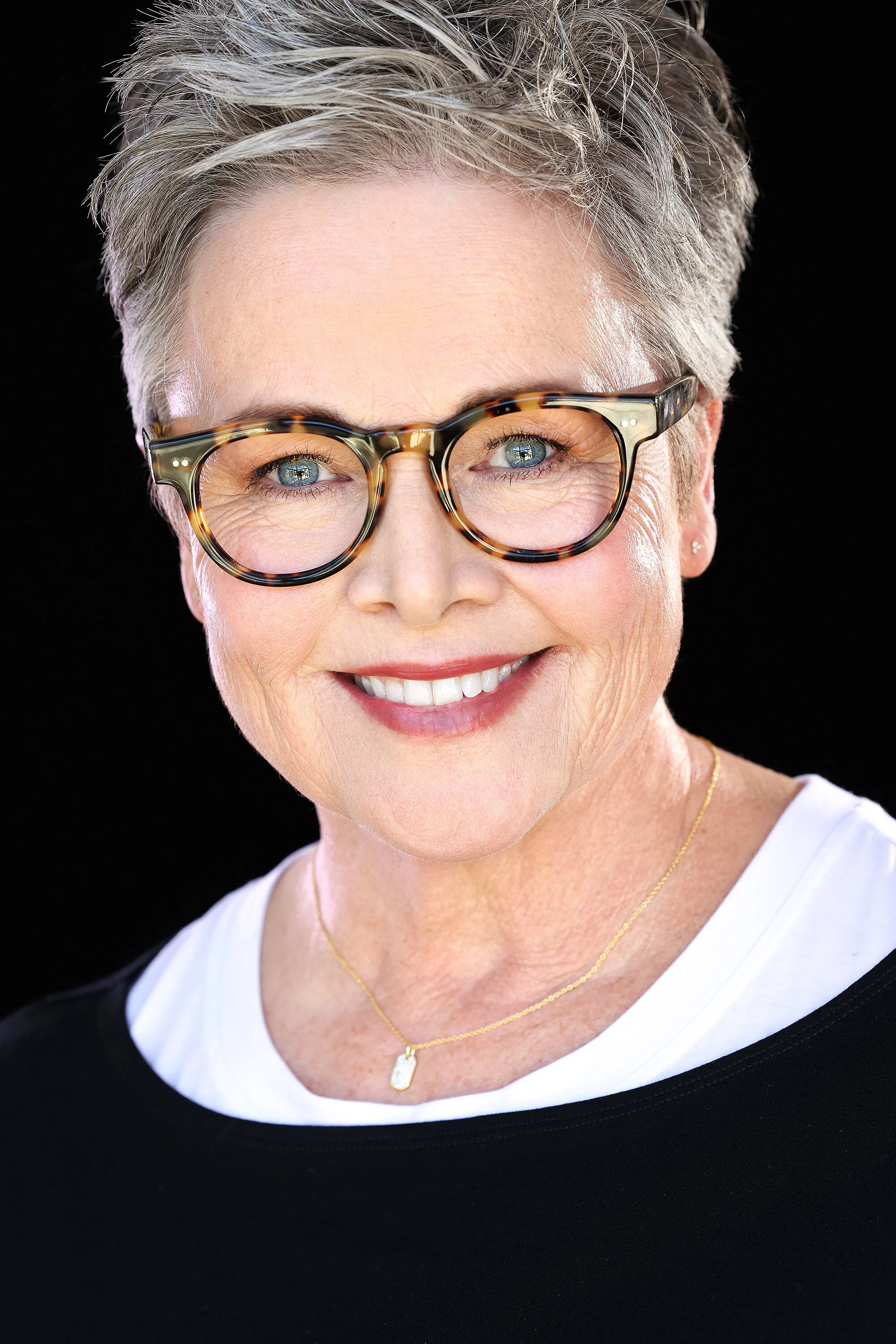 A smiling older woman with short gray hair, wearing tortoiseshell glasses, a gold necklace, and a black top with a white neckline, against a black background.