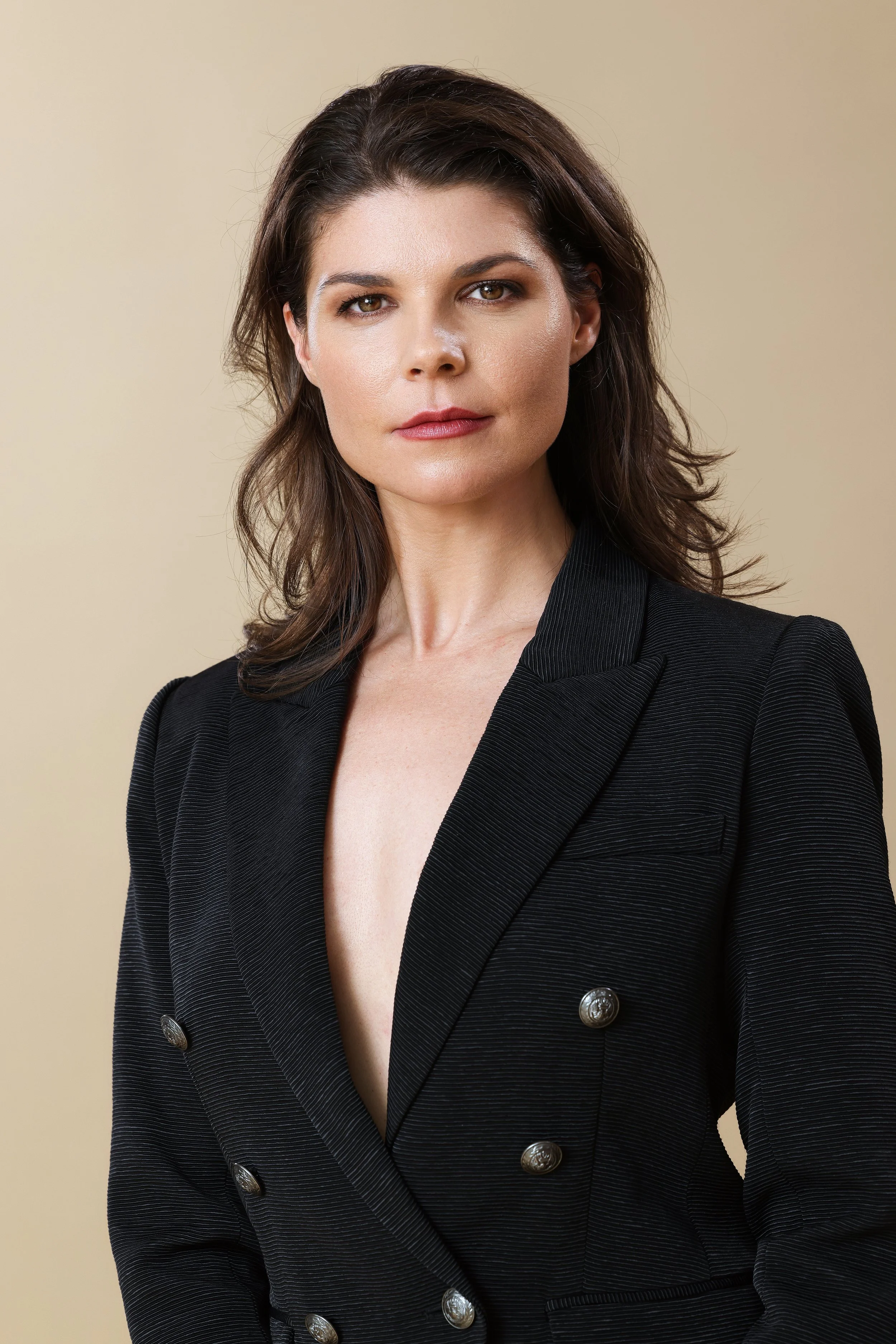 A woman with shoulder-length brown hair wearing a black blazer with silver buttons, standing against a beige background.