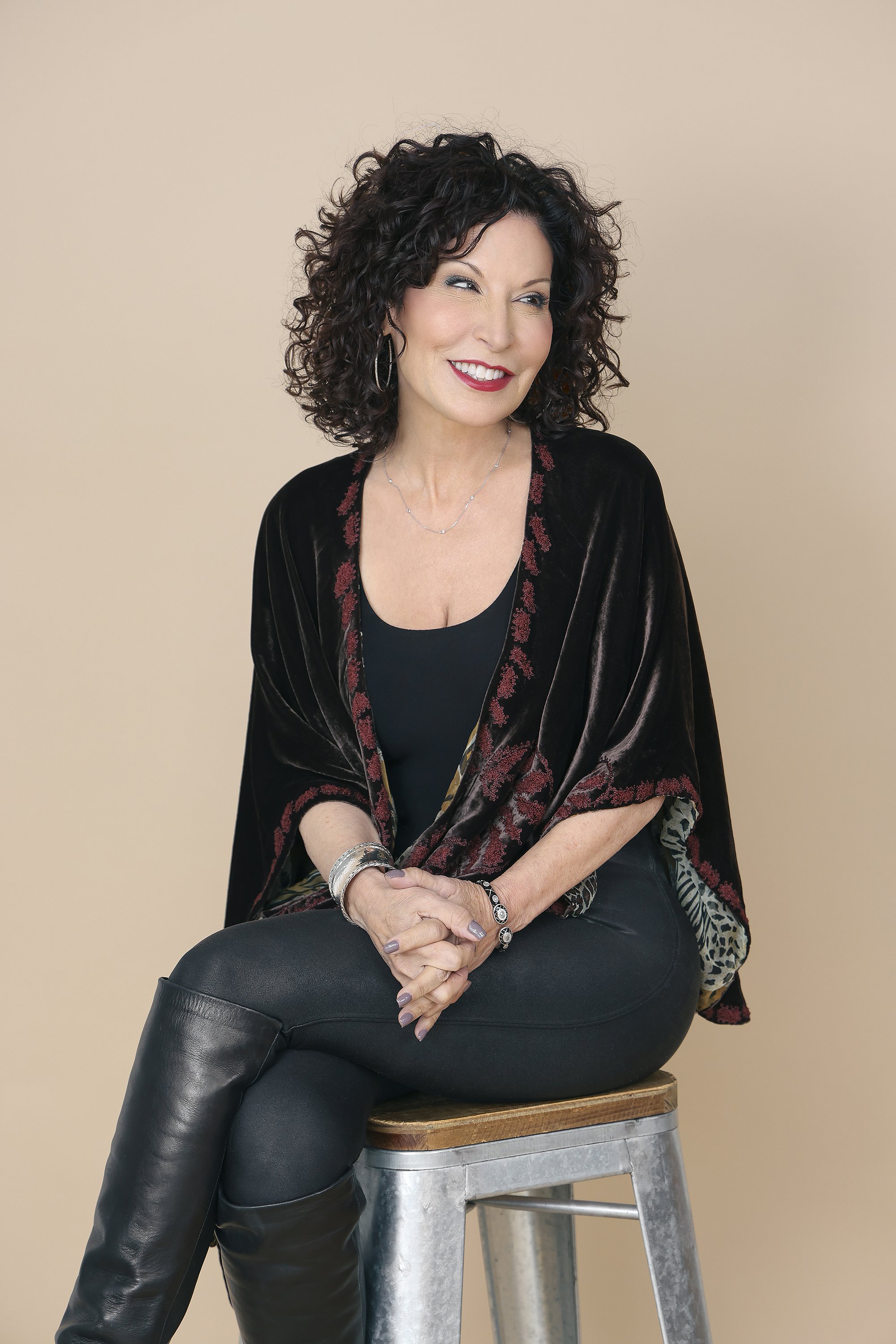 A woman with dark, curly hair sitting on a stool against a beige background. She is wearing a black top, black leather pants, and a dark velvet shawl with floral embroidery. She has jewelry including bracelets, rings, and earrings, and is smiling whi