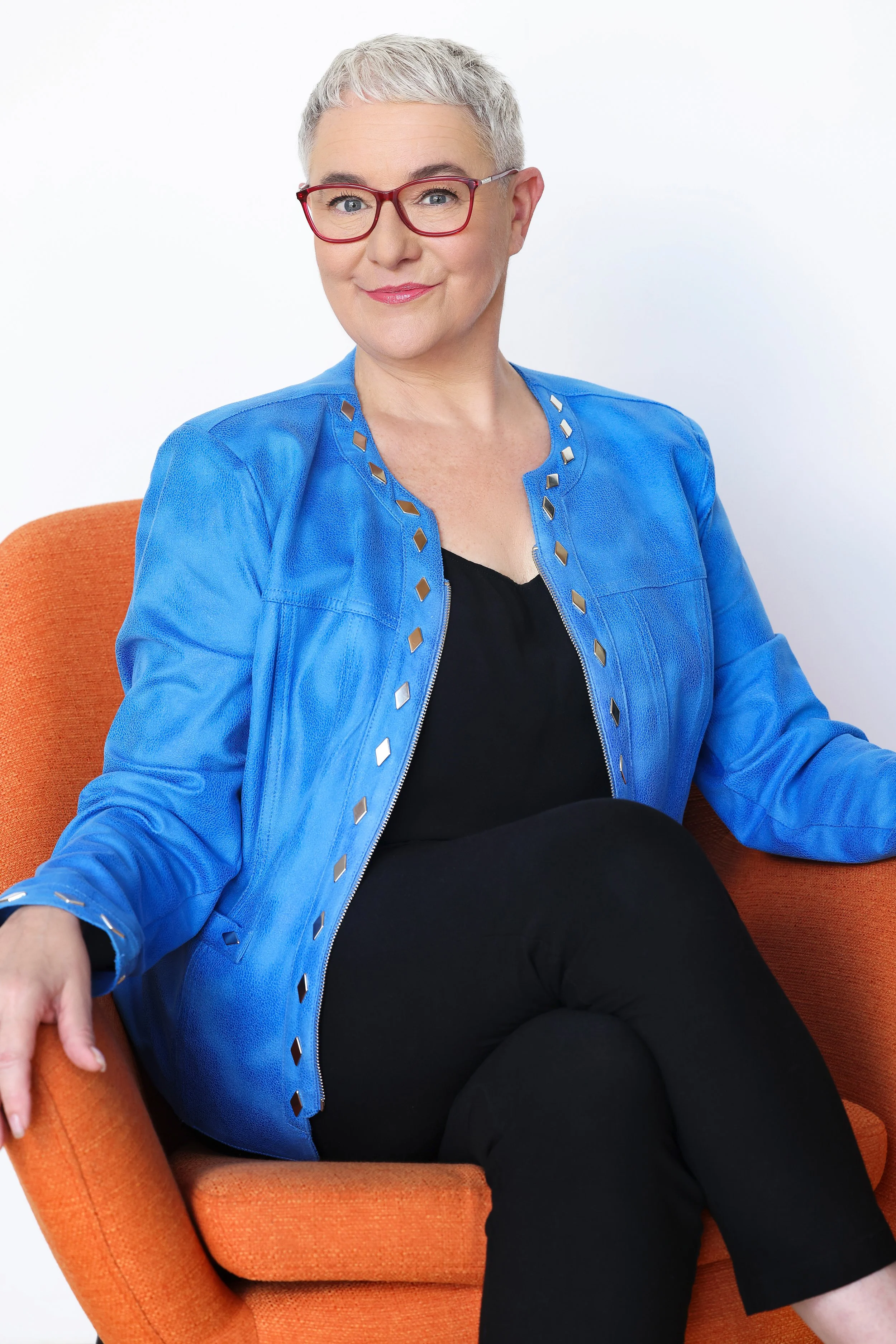A woman with short gray hair and red glasses sitting in an orange armchair, wearing a bright blue jacket, a black top, and black pants, smiling at the camera against a plain white background.