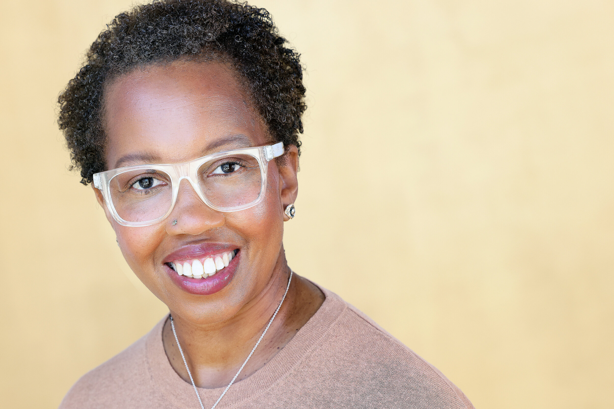 A smiling woman with short curly hair, wearing clear glasses, a nose piercing, earrings, a necklace, and a beige top, stands against a yellow background.