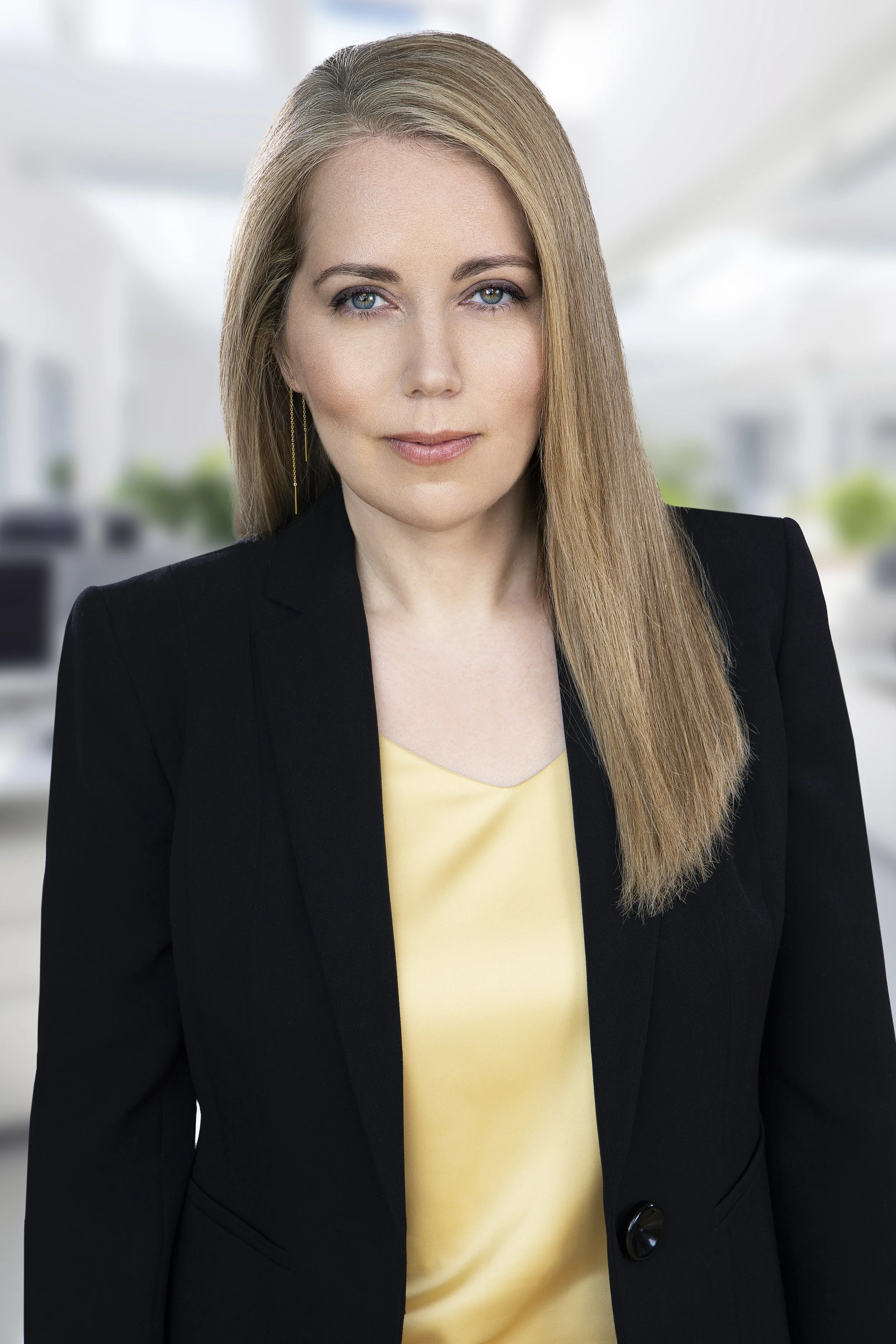 A professional woman with straight blonde hair, blue eyes, and light makeup, wearing a black blazer and a yellow blouse, standing in a bright office environment.