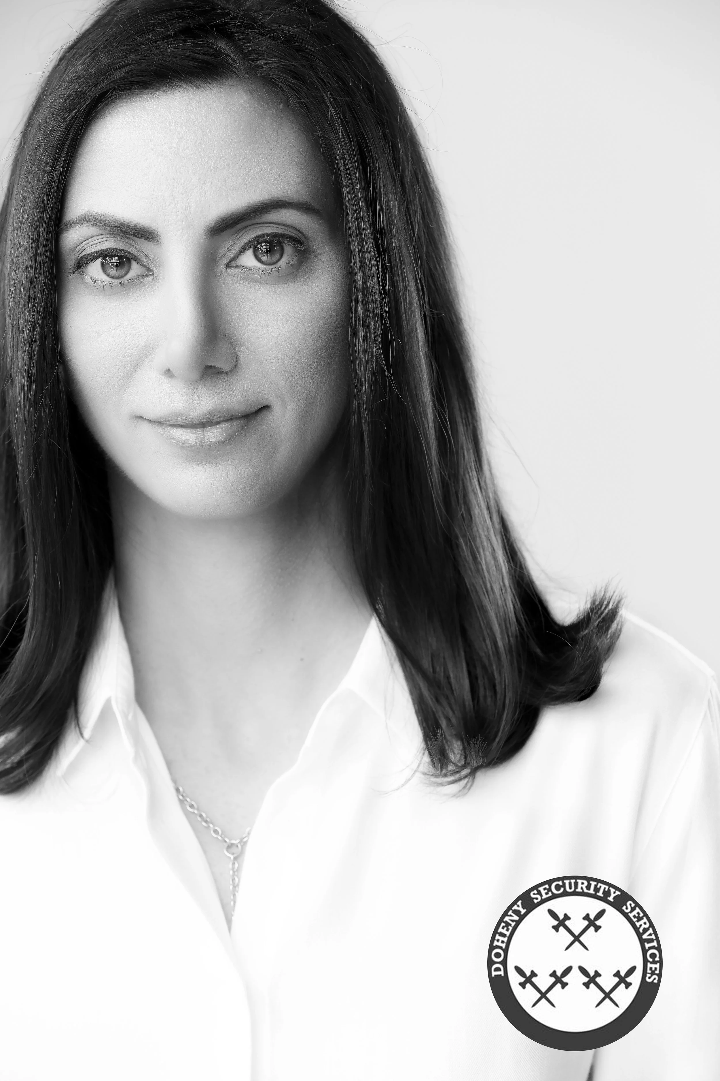 Black and white portrait of a woman with long dark hair, wearing a white shirt with an embroidered patch that says "Doheny Security Services" with crossed tools inside a circle.