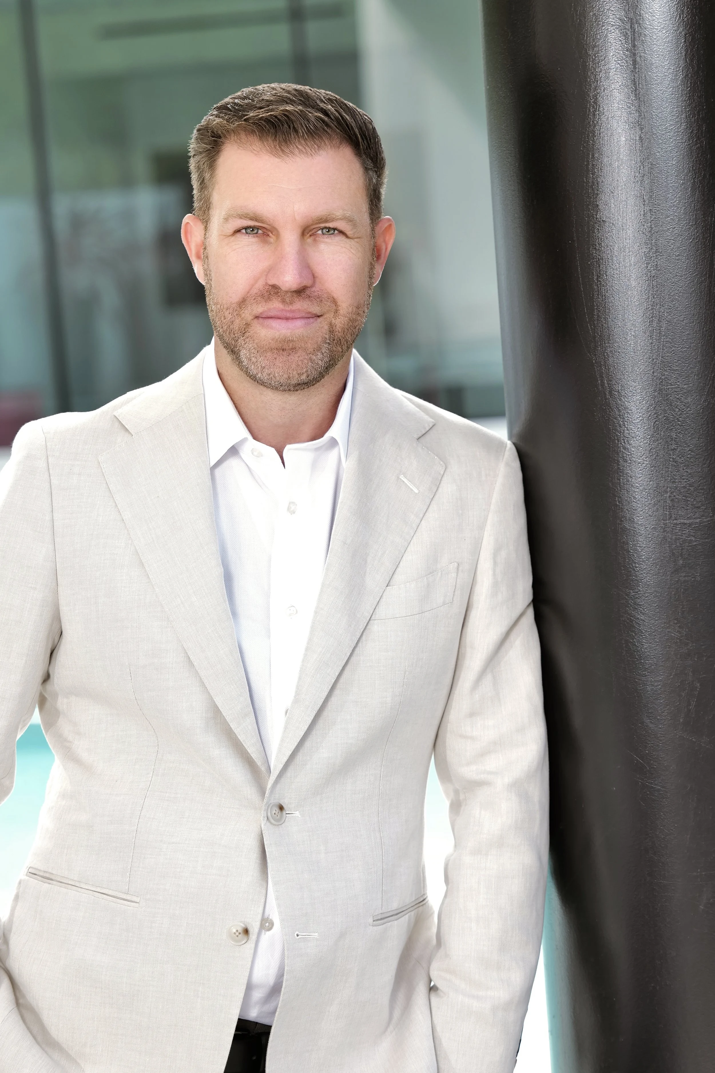 A man with short brown hair, a beard, and blue eyes wearing a light beige suit jacket and white shirt, standing outdoors near a black column with a cityscape background.