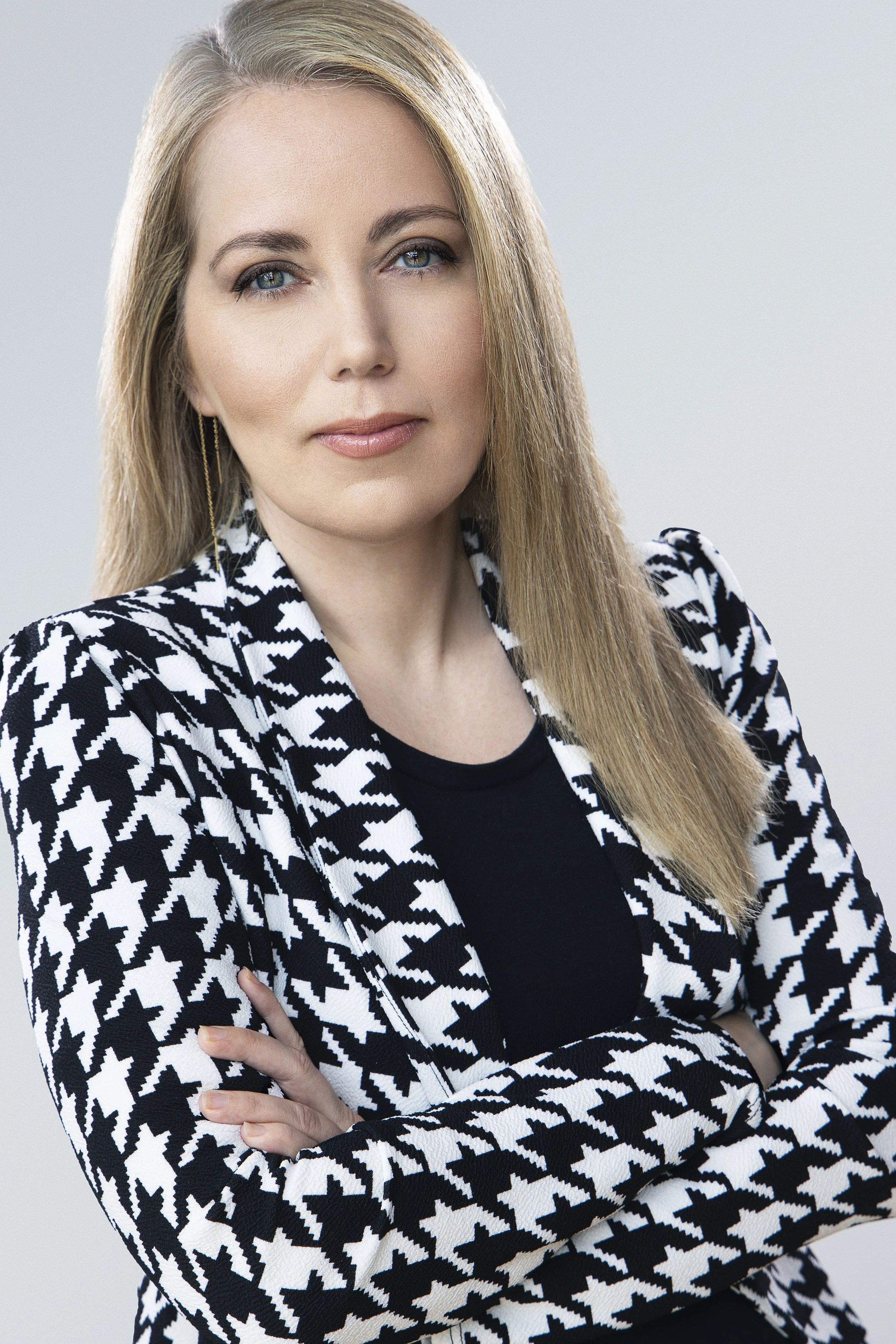A woman with long blonde hair and blue eyes wearing a black top and a black and white houndstooth patterned blazer, crossing her arms, against a plain light gray background.