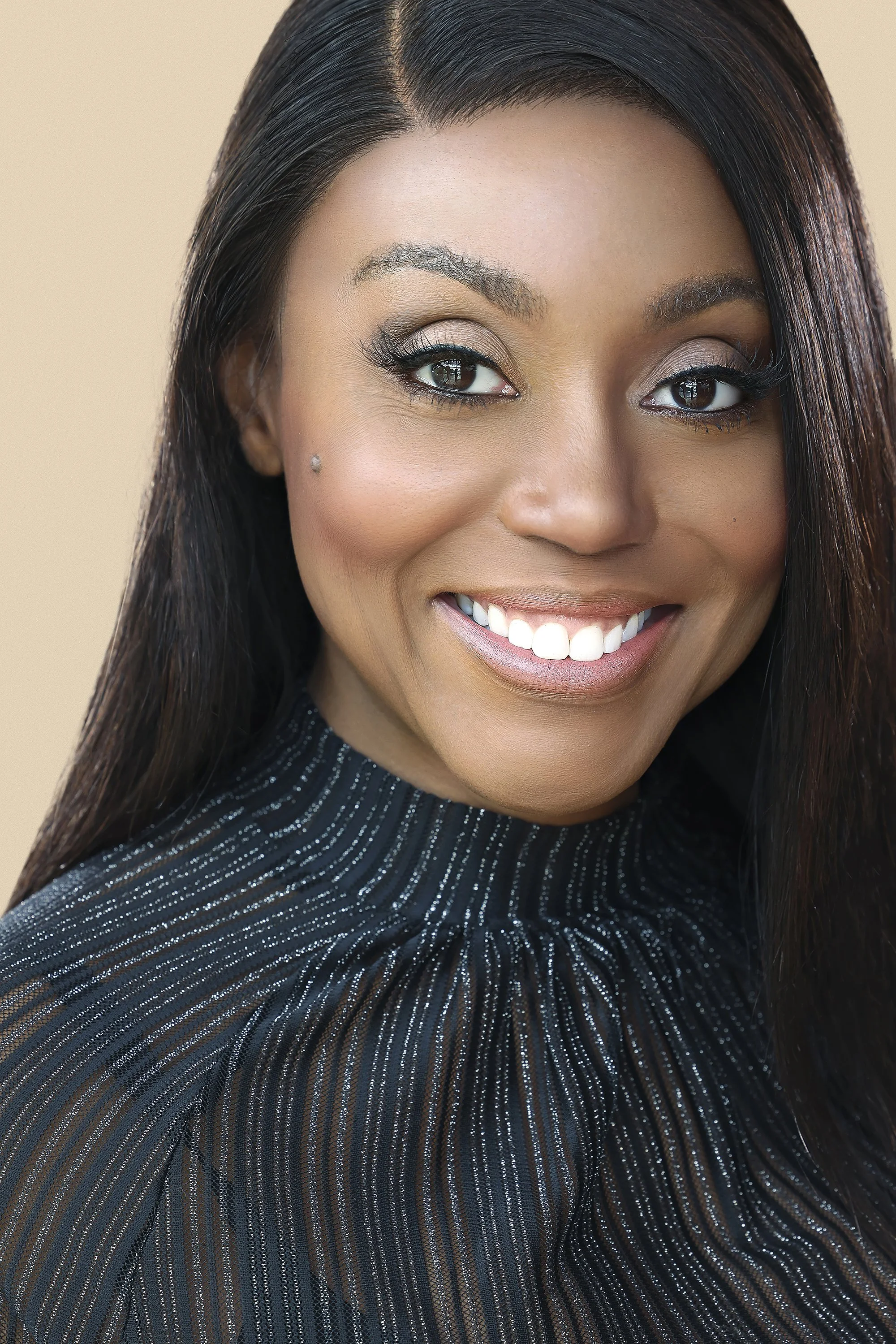 Close-up portrait of a smiling woman with long dark hair, light makeup, and a small diamond earring, wearing a dark, sparkly, high-neck blouse against a beige background.