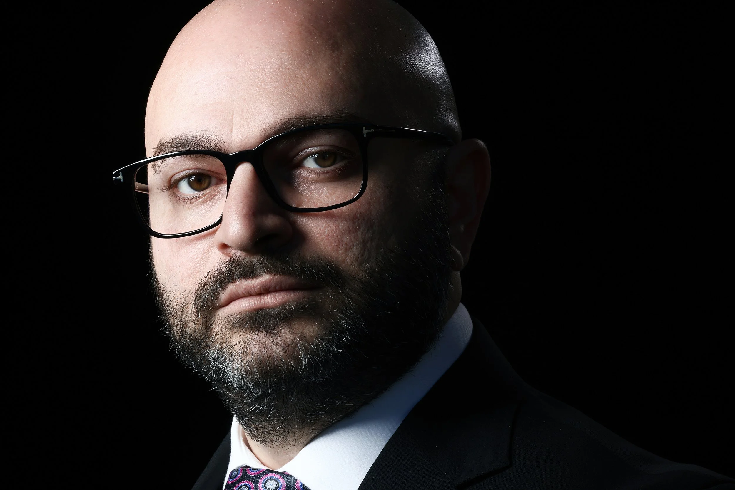 A portrait of a bald man with a beard wearing black glasses and a suit, set against a dark background.