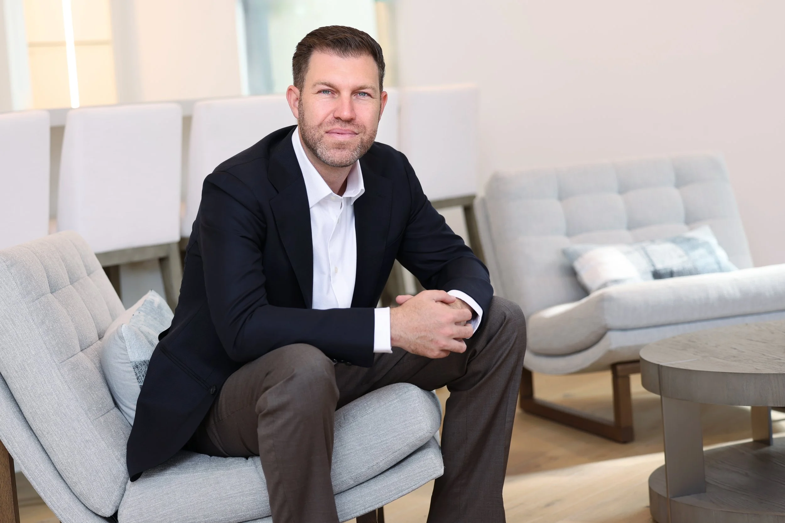 A man in a dark blazer and white shirt sitting on a light-colored sofa in a modern living room, looking at the camera.