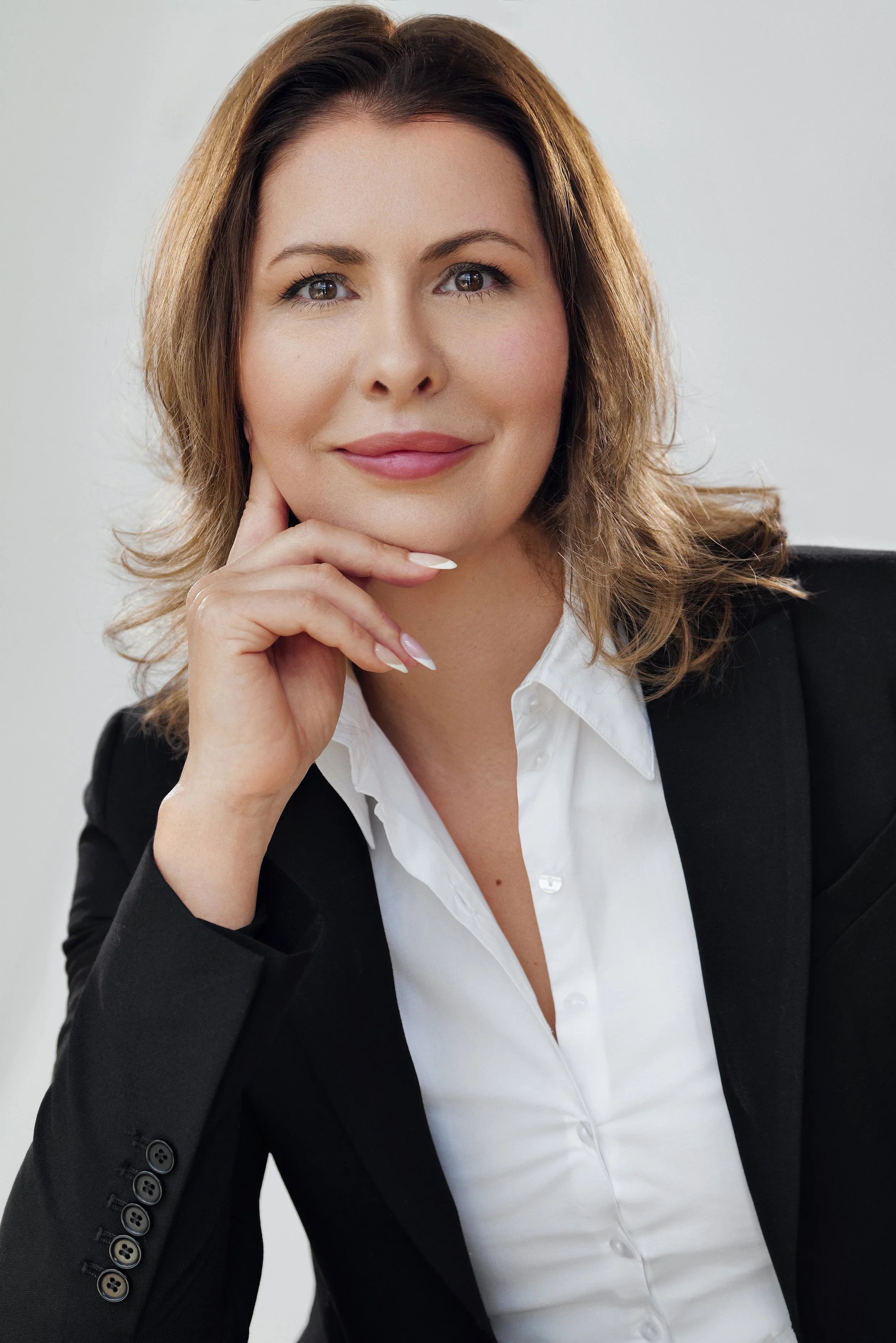 A portrait of a confident woman with shoulder-length, wavy brown hair, wearing a black blazer and white shirt, resting her chin on her hand, looking directly at the camera with a slight smile.