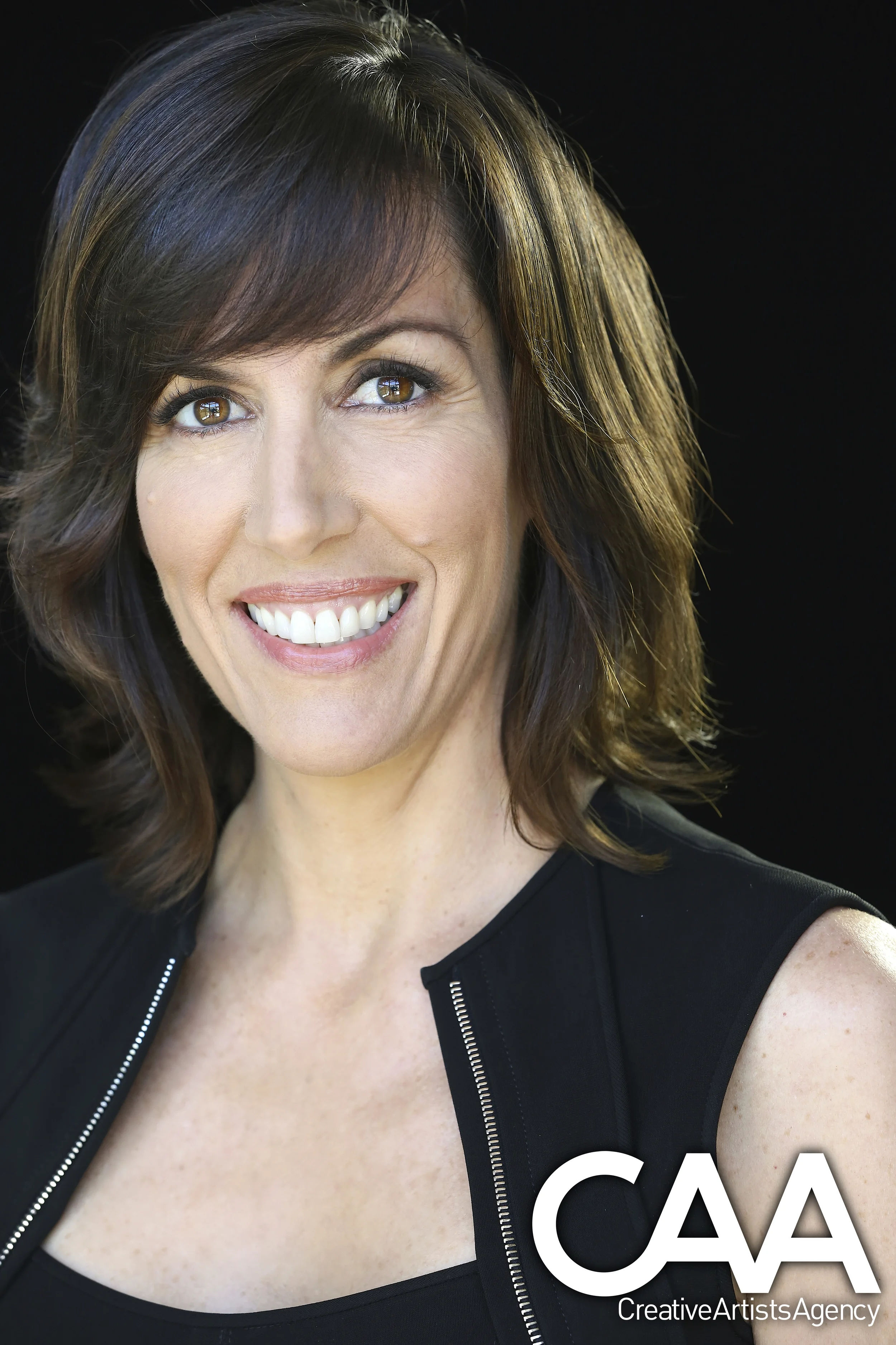 Close-up portrait of a smiling woman with brown hair, wearing a black top and vest, against a dark background.