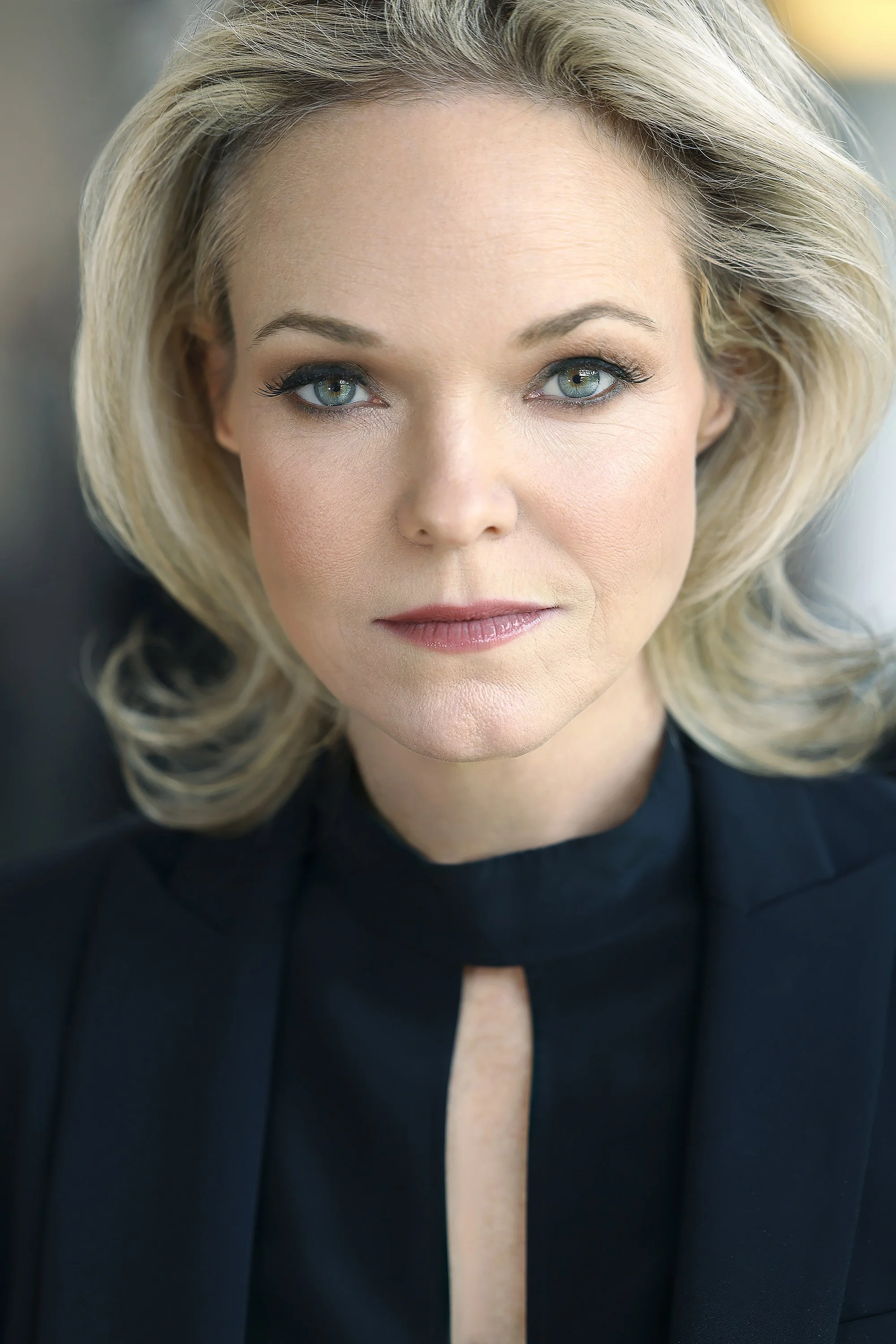 Close-up of a woman with blonde hair, blue eyes, wearing a black top, looking directly at the camera.