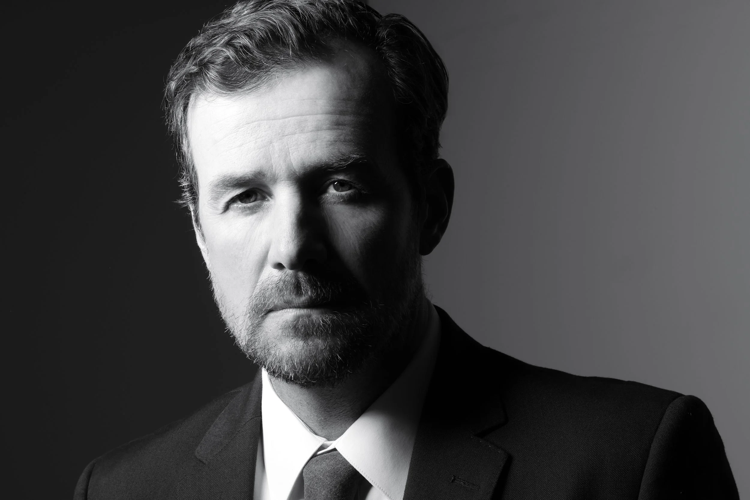 Black-and-white portrait of a man with a beard and styled hair, wearing a suit and tie, looking directly at the camera.
