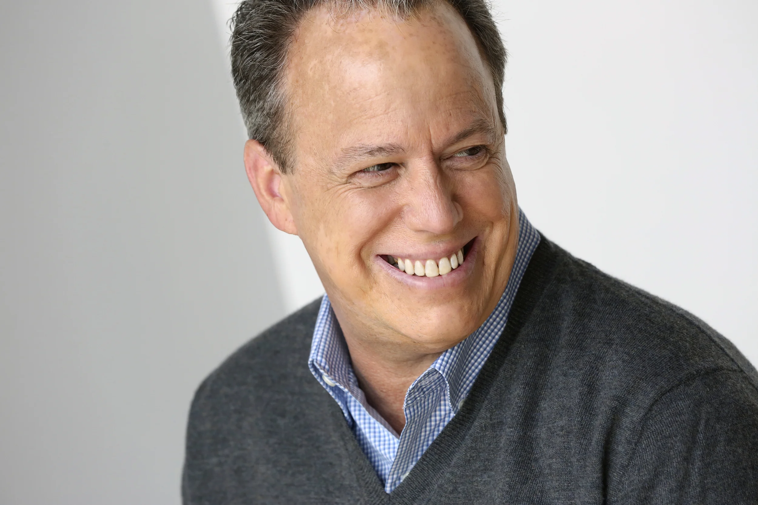 A middle-aged man smiling while looking to his left. He has short, dark hair and is wearing a blue checkered shirt with a dark gray sweater over it.