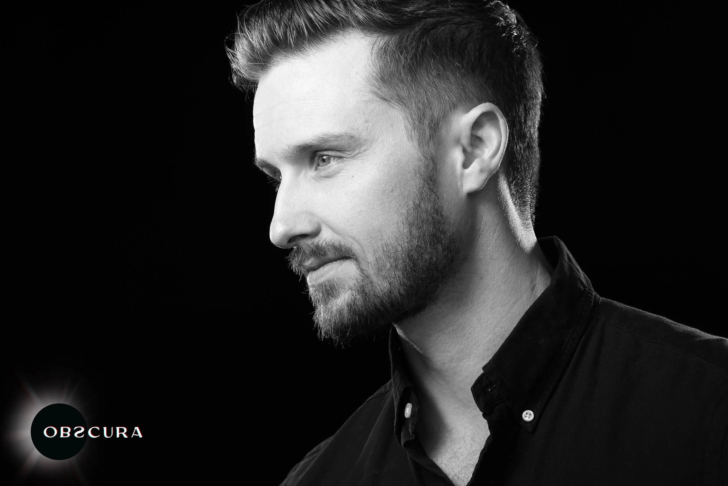 Black and white portrait of a man with short styled hair and a beard, looking to the side with a neutral expression, against a black background, with the word 'OBSCURA' in the lower left corner.