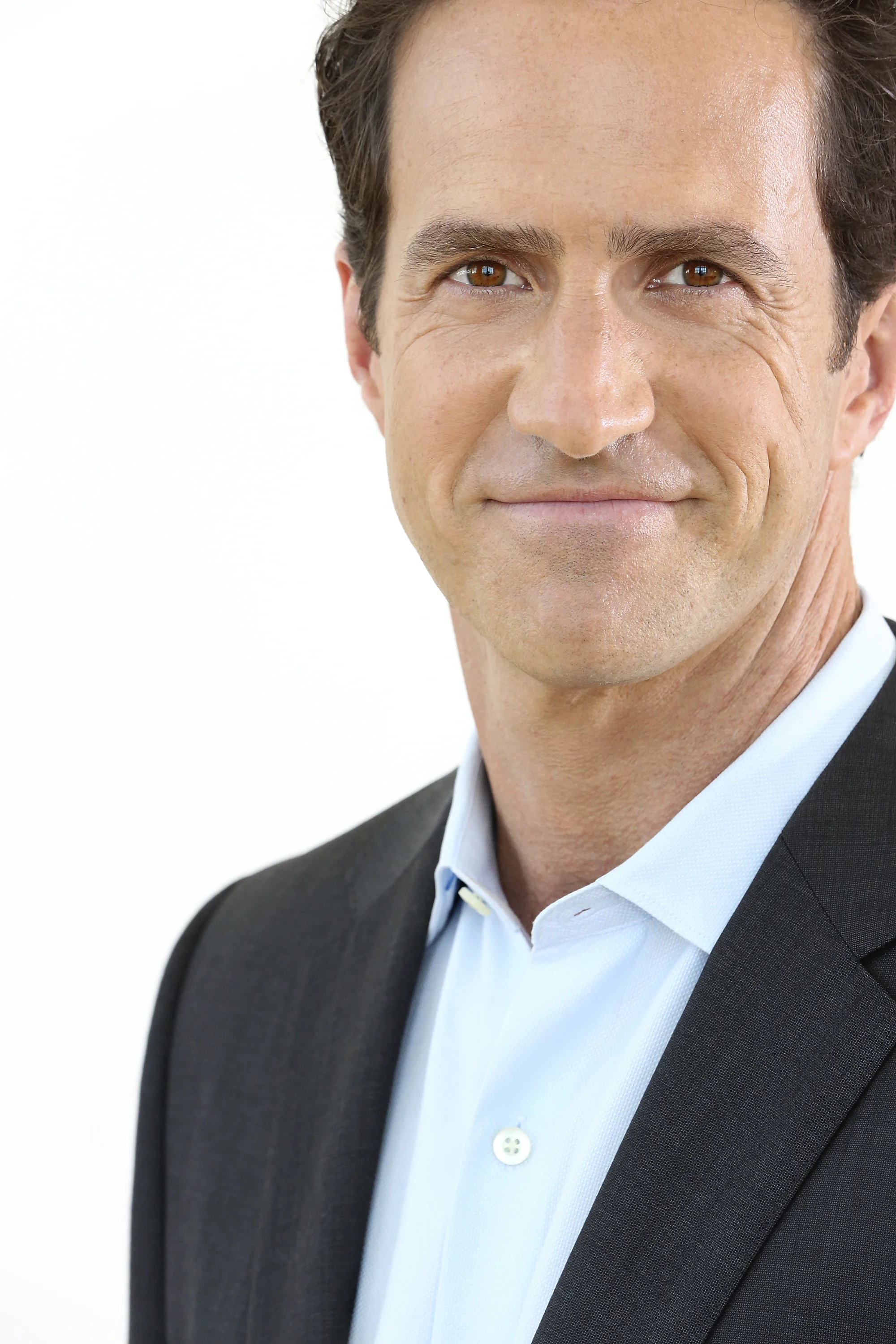 A middle-aged man with brown hair and light skin, smiling, wearing a light blue dress shirt and a black suit jacket, standing against a white background.