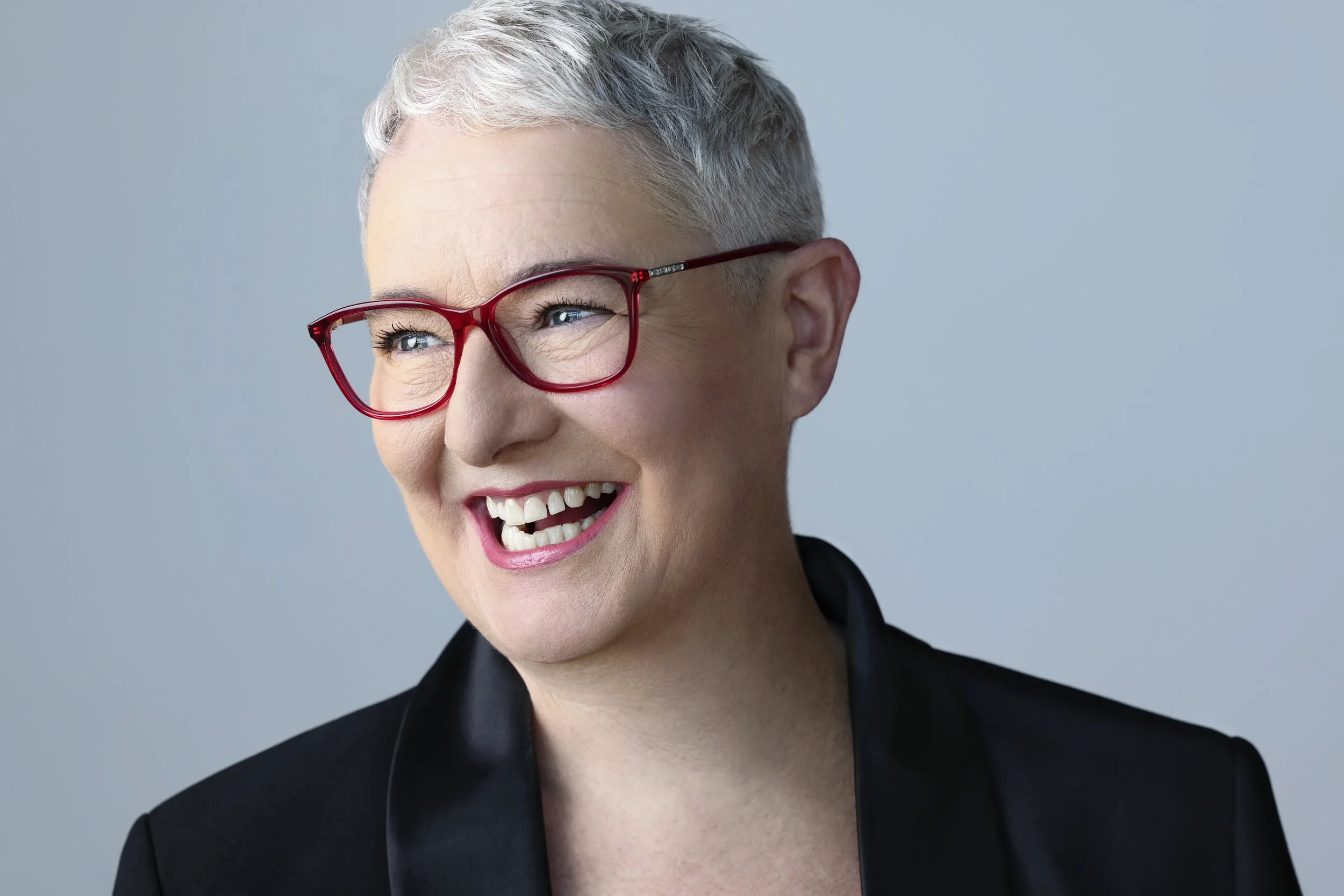 A smiling woman with short gray hair, wearing red glasses and a black blazer, looking to the side. She appears to be happy or amused.