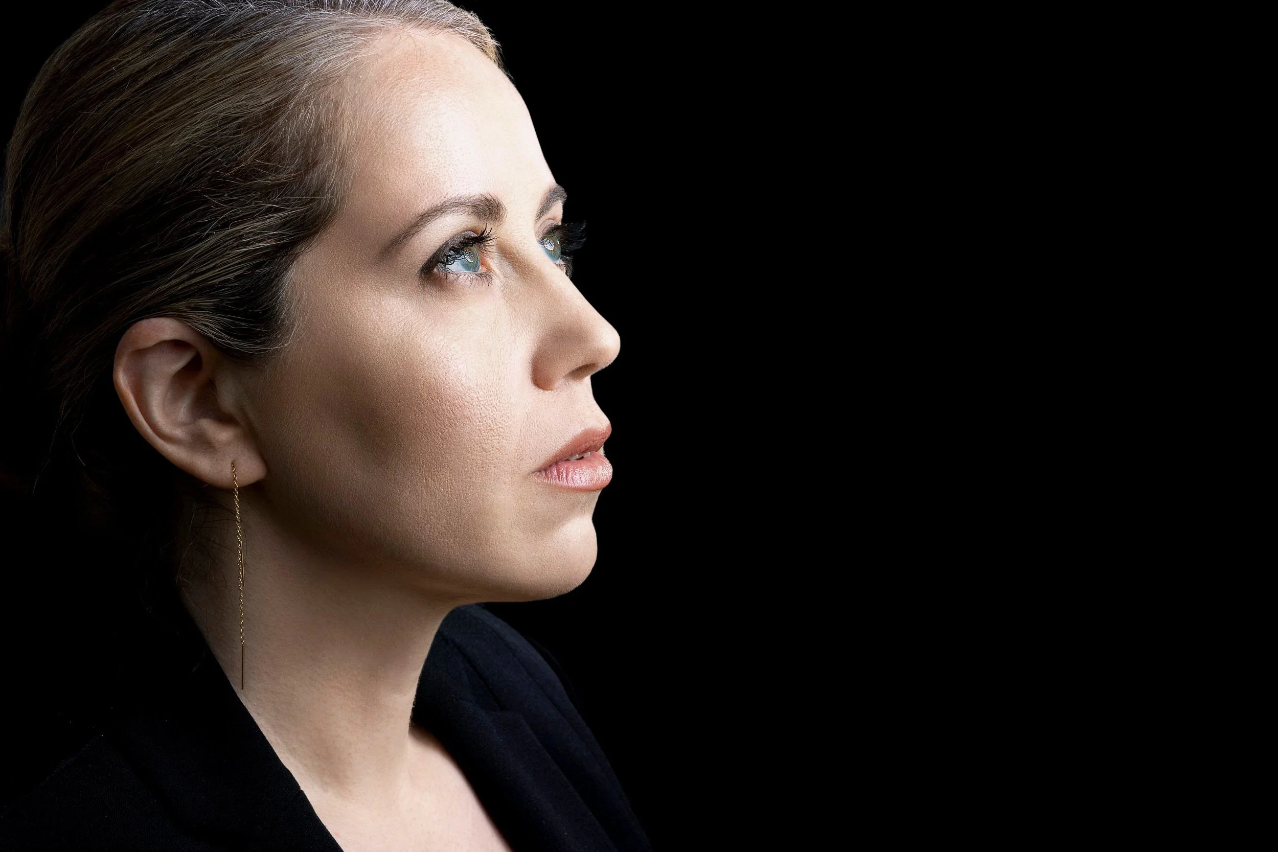 Close-up profile of a woman with fair skin and blue eyes, wearing an earring with a black background.