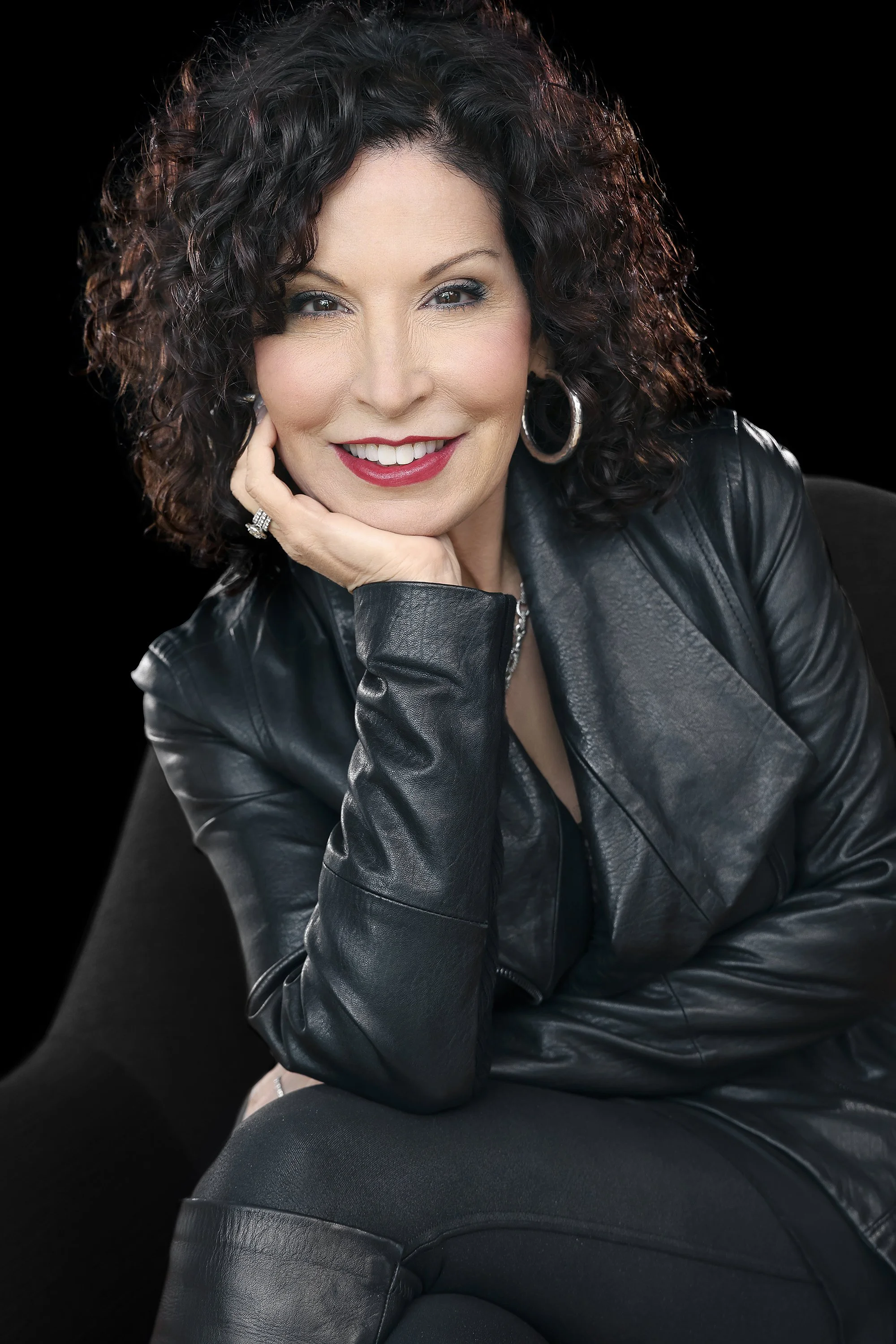 A woman with curly dark hair smiling, wearing a black leather jacket, silver hoop earrings, and a ring, resting her chin on her hand, against a black background.