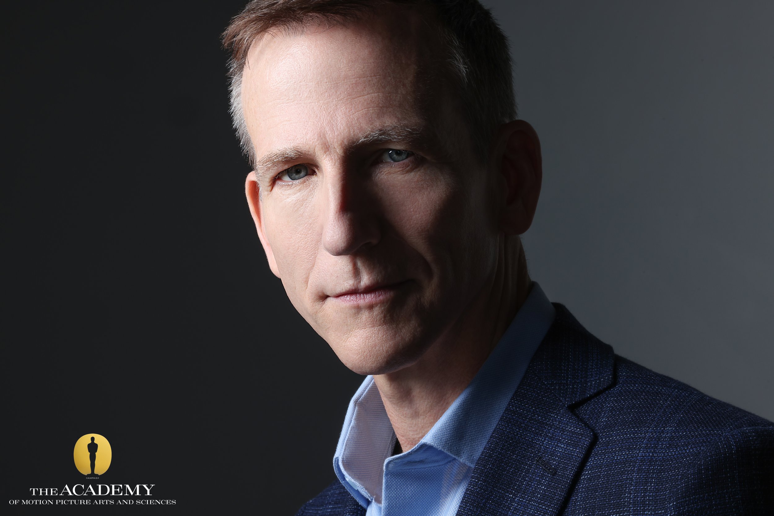 A professional headshot of a middle-aged man with short brown hair, blue eyes, wearing a blue suit and light blue shirt, against a dark gray background. Logo of The Academy of Motion Picture Arts and Sciences is in the bottom left corner.