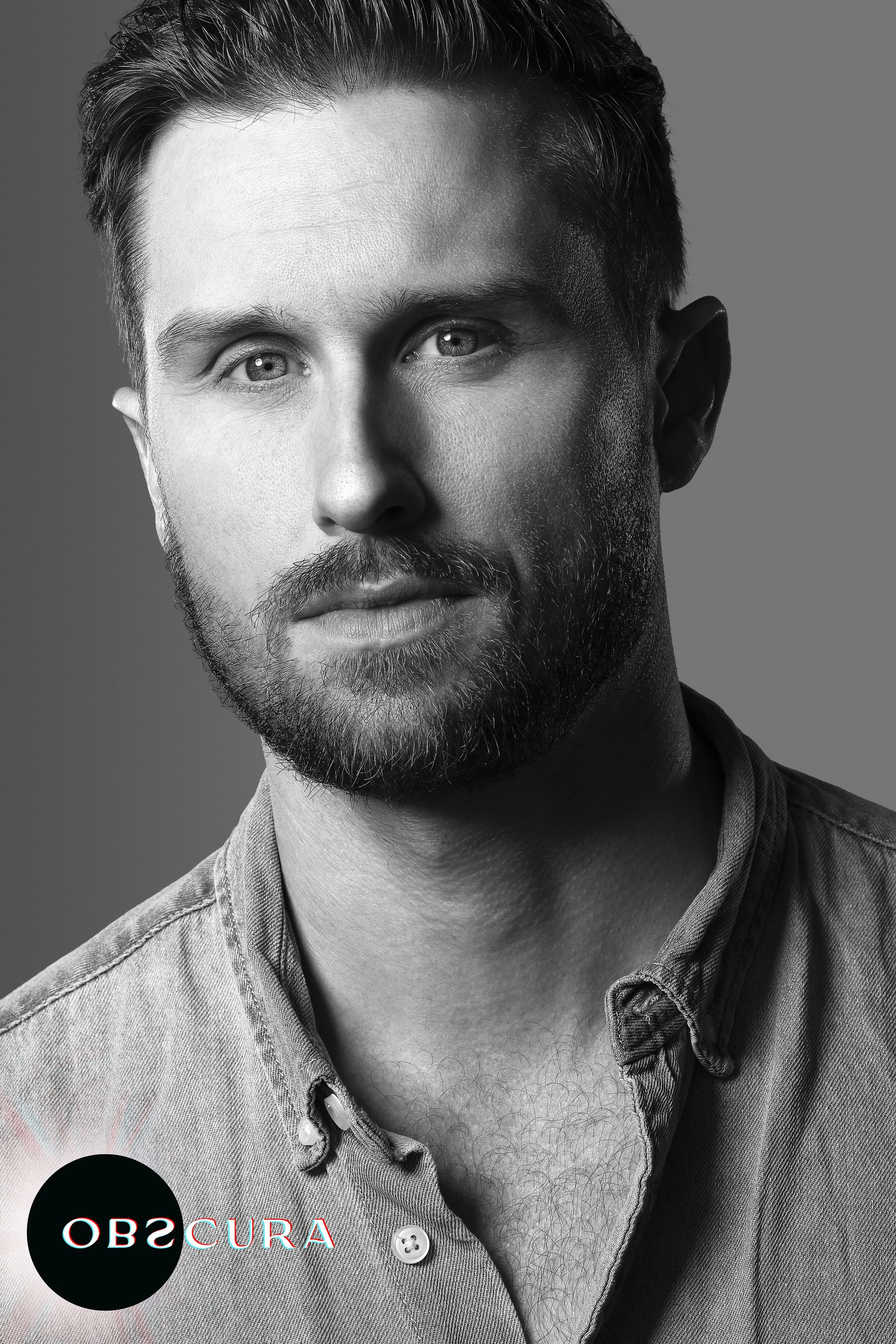 Black-and-white portrait of a man with short hair, a beard, and mustache, wearing a button-up shirt with the top button undone, looking directly at the camera with a neutral expression.
