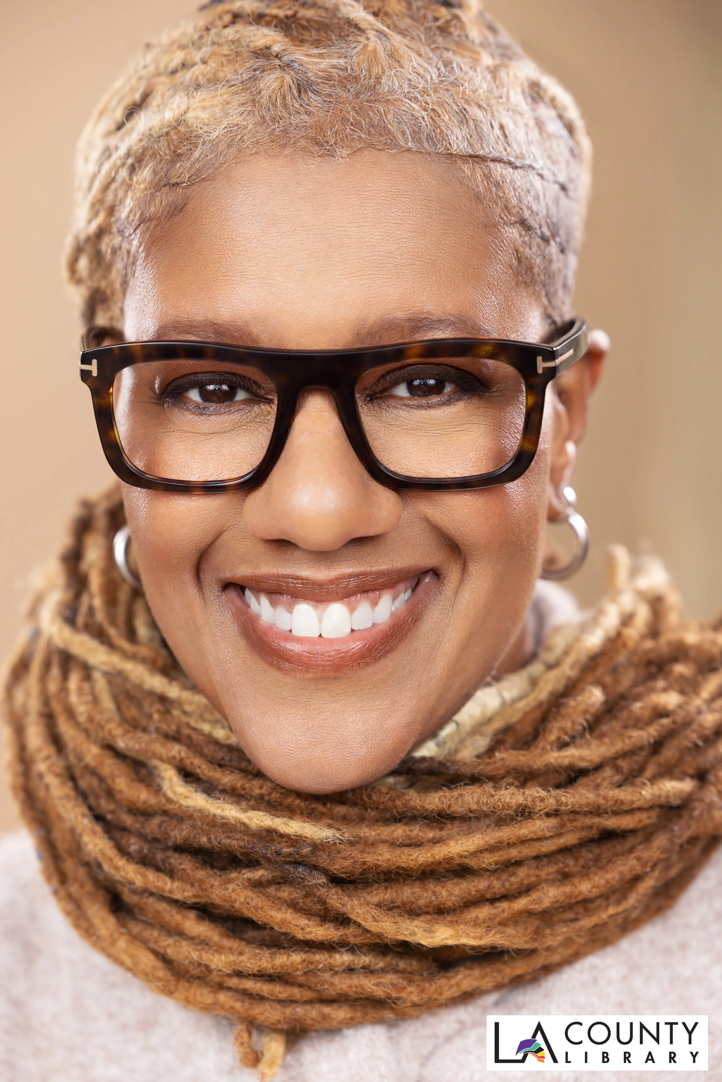 A smiling woman with short, blonde dreadlocks, wearing black and tortoise-shell glasses, hoop earrings, and a knitted scarf.