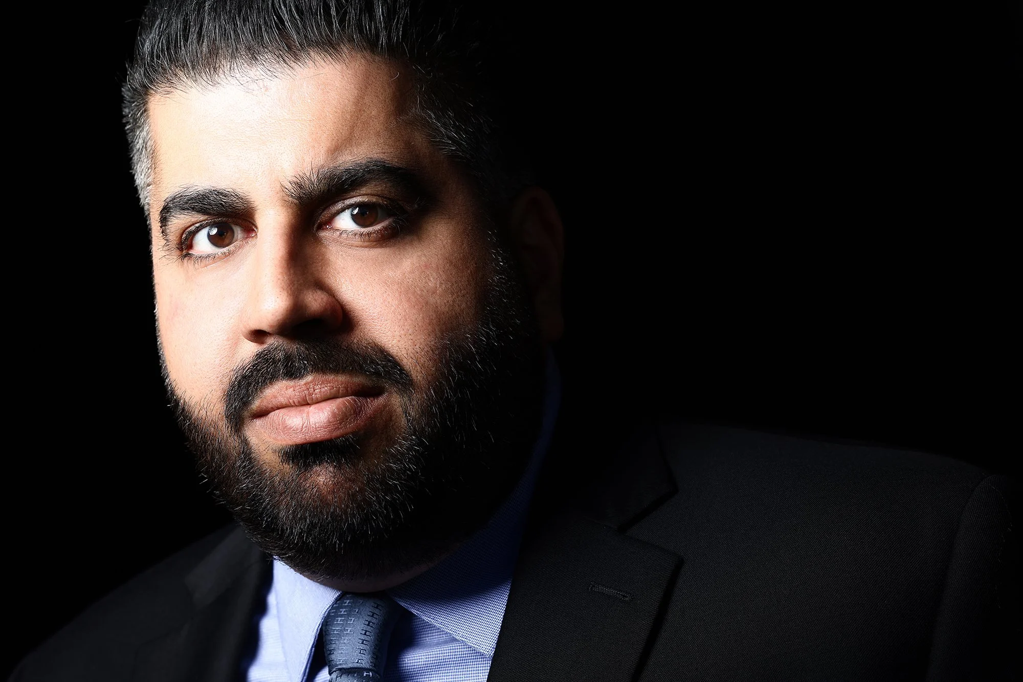 Close-up portrait of a man with dark hair, beard, and mustache, wearing a suit and tie, against a black background