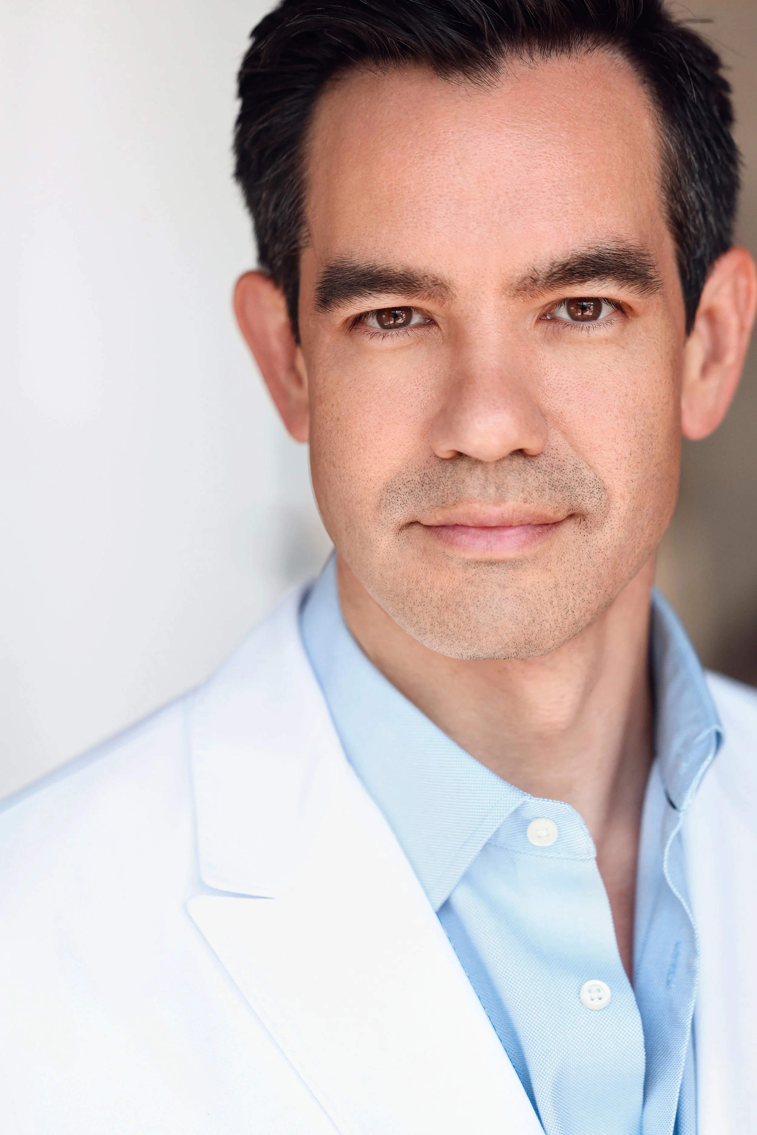 Close-up portrait of a man in a light blue collared shirt and a white blazer, smiling subtly with dark hair and brown eyes, against a plain background.
