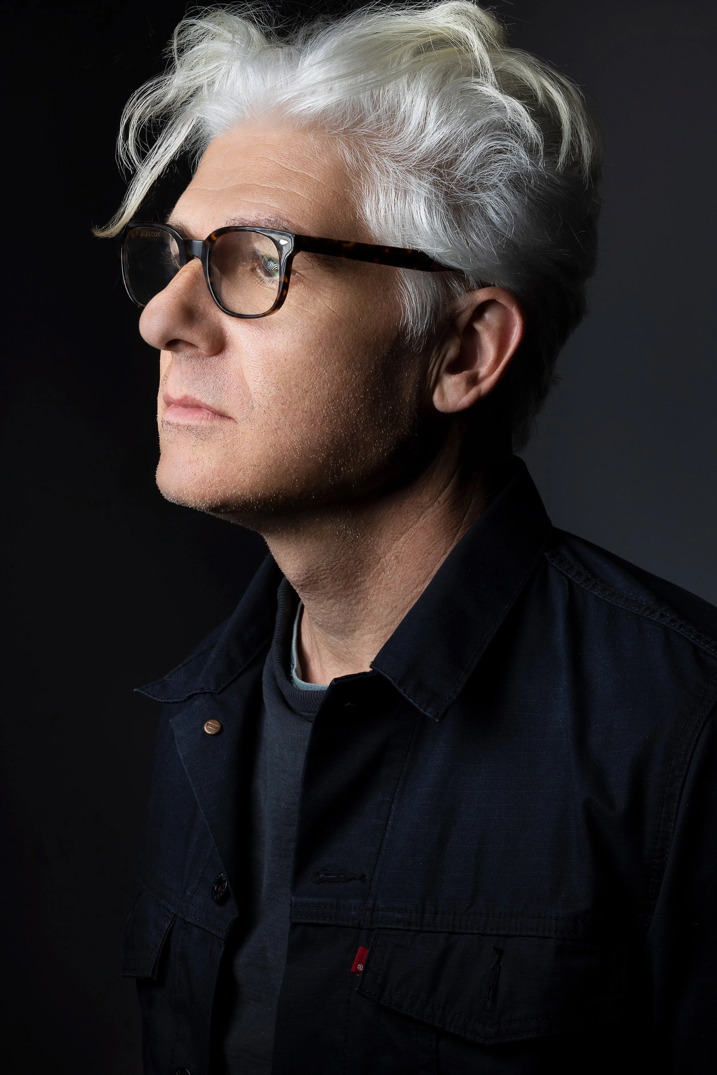 A side profile portrait of a middle-aged man with white hair, glasses, and light skin, wearing a black shirt against a dark background.