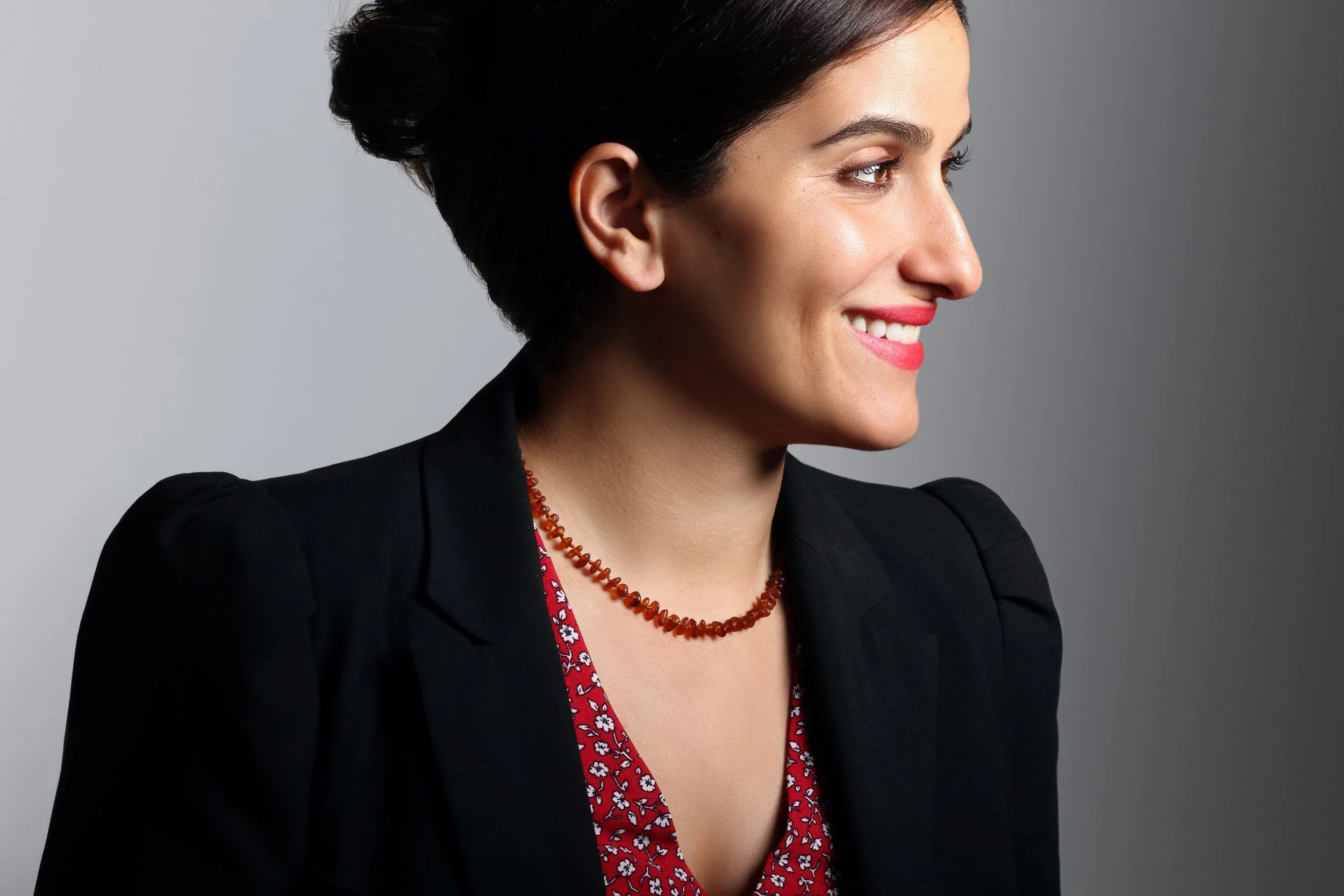 Side profile of a woman with short black hair, wearing a black blazer, red floral blouse, and red beaded necklace, smiling against a gray background.