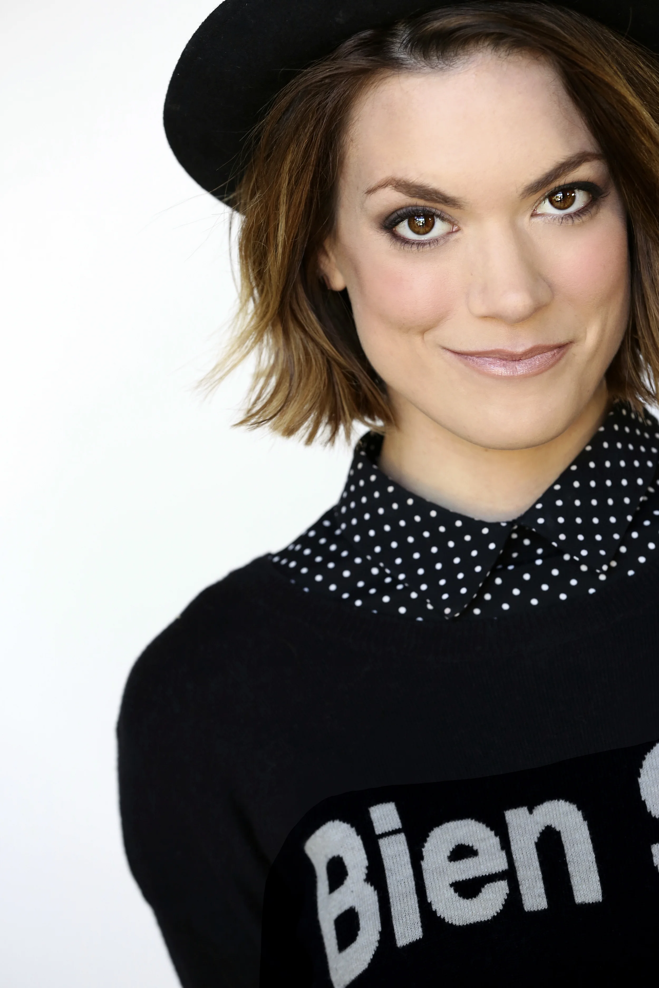 A woman with shoulder-length brown hair wearing a black hat, a black sweater with the word 'Bien' partially visible, and a polka dot shirt underneath, smiling softly against a white background.