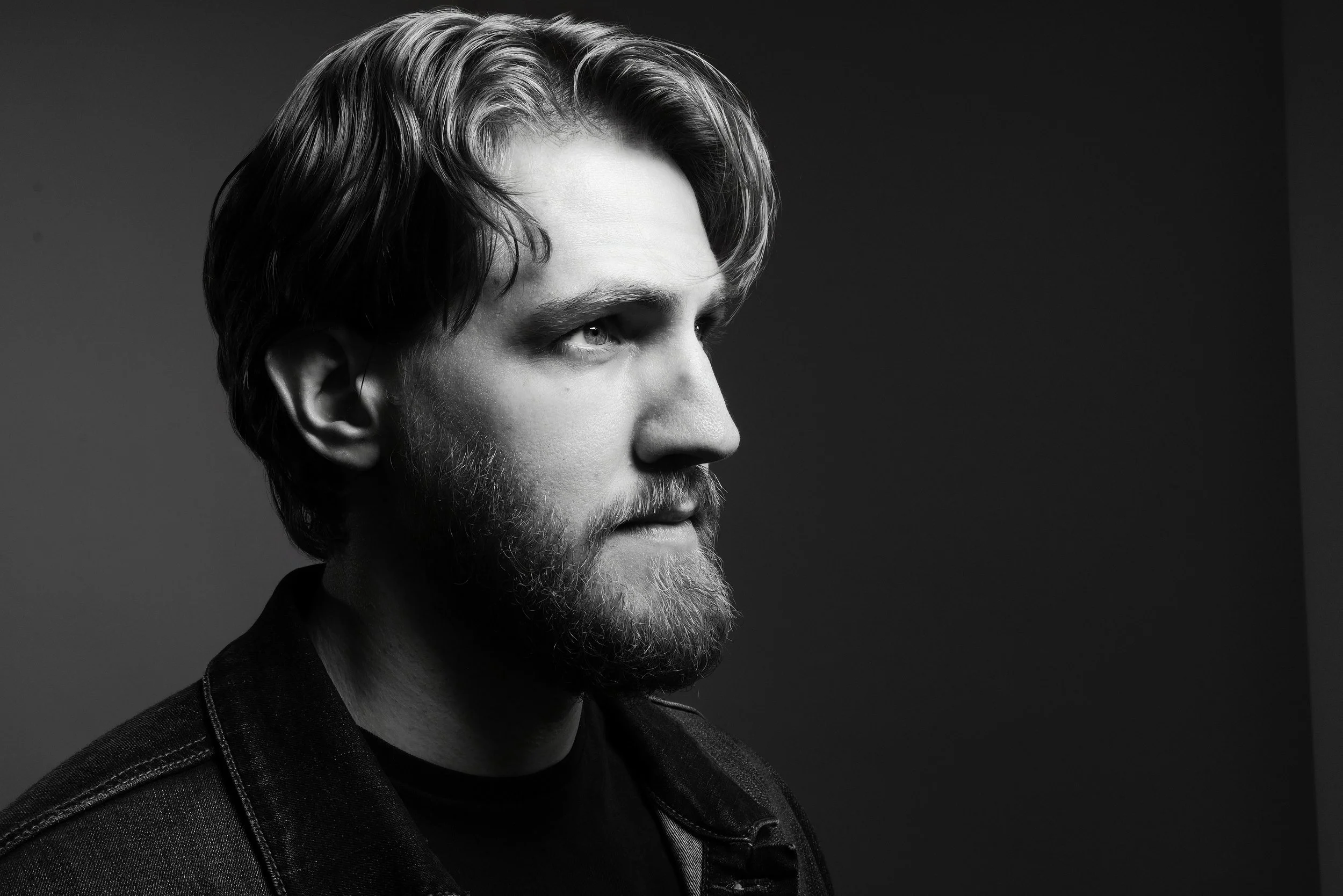 A black-and-white portrait of a man with a beard and wavy hair, looking to the right, wearing a dark jacket, with a plain dark background.