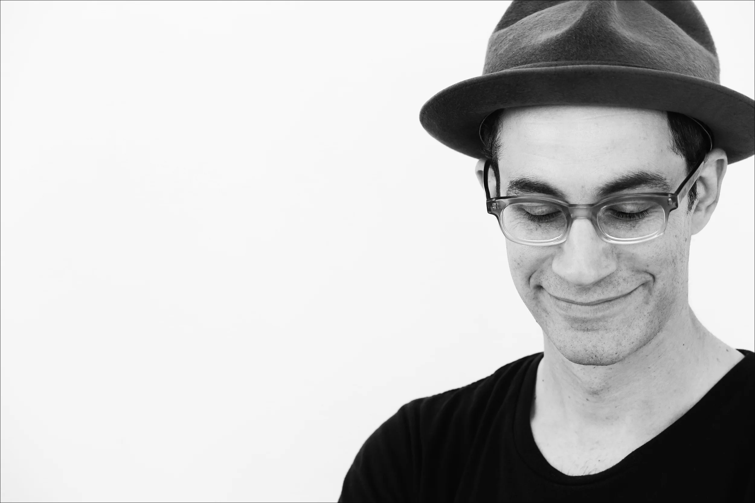 Black and white photo of a smiling man wearing glasses and a fedora hat, looking down.