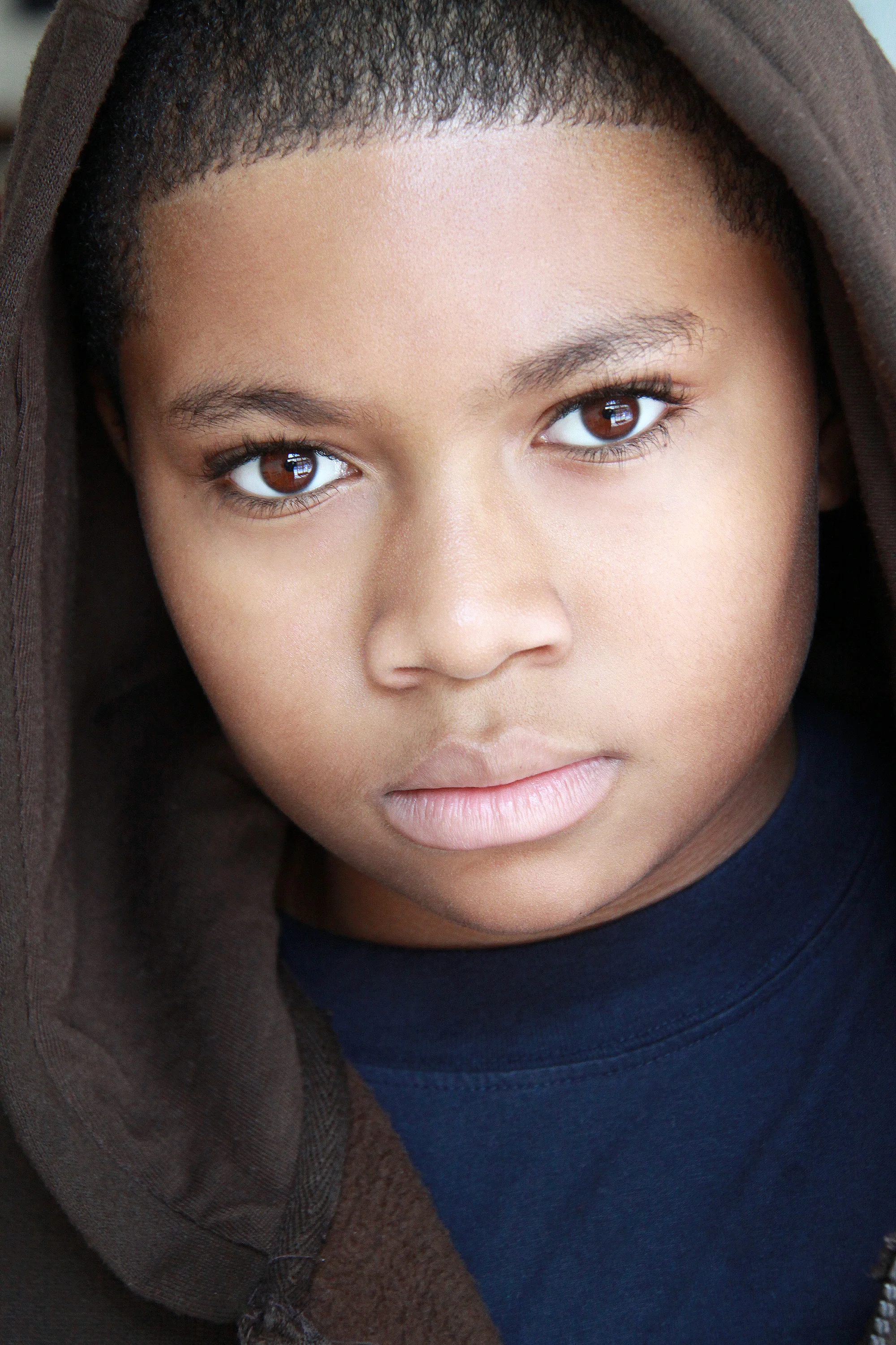 Close-up of a young African American boy with short hair, brown eyes, wearing a dark hoodie and navy blue shirt.