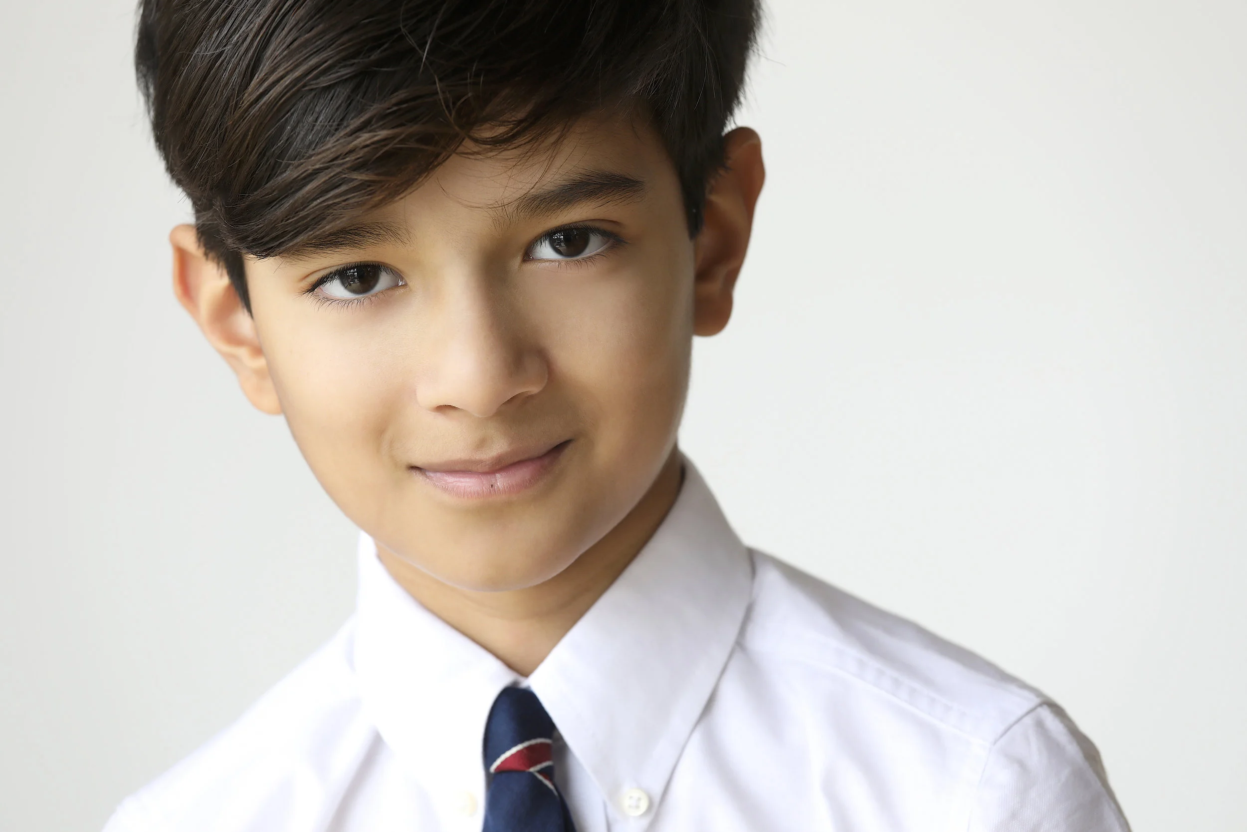 Young boy with dark hair wearing a white shirt and tie, smiling softly against a plain background.