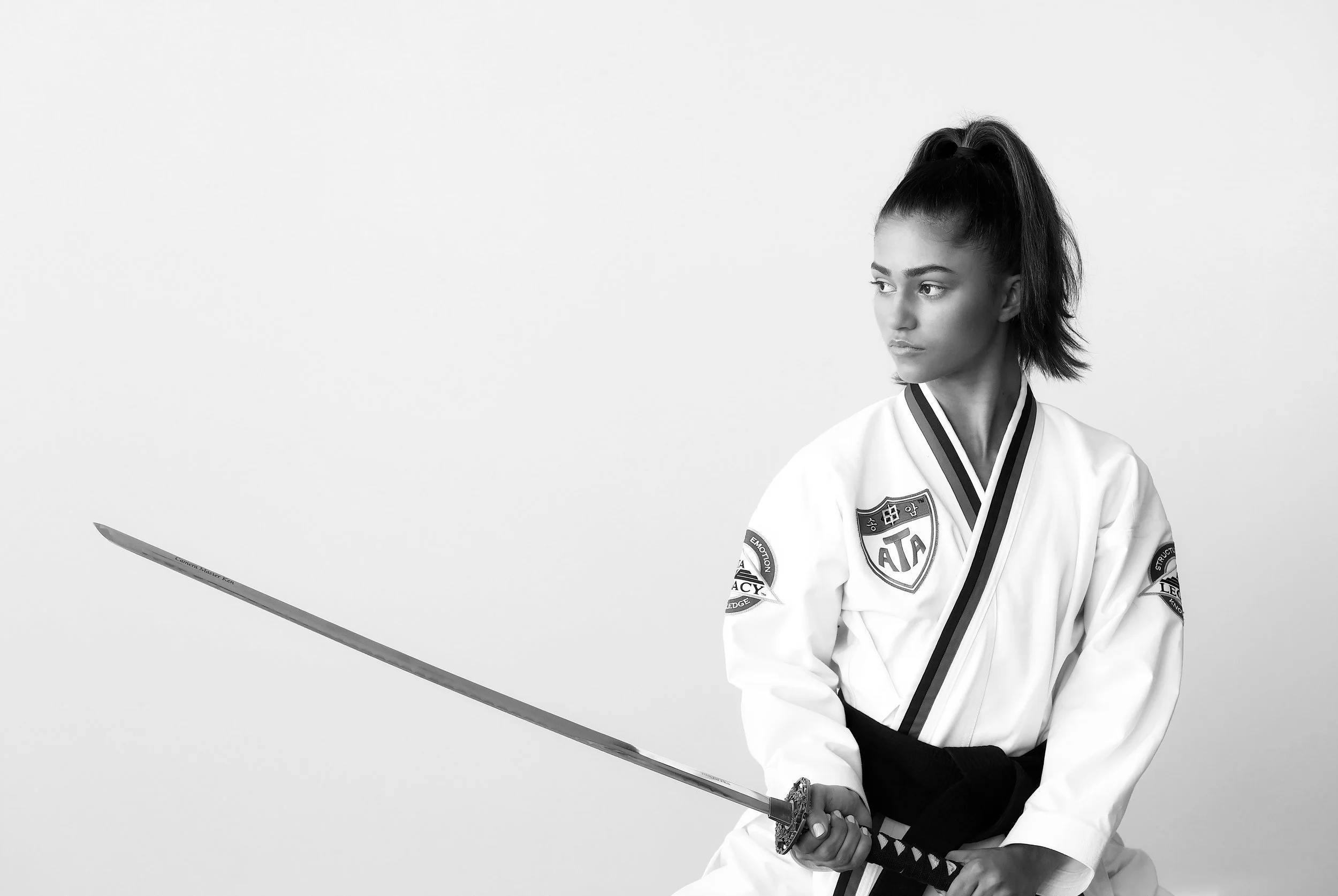 Black and white photo of a woman in a martial arts uniform holding a sword, sitting in a traditional pose, looking to the right.