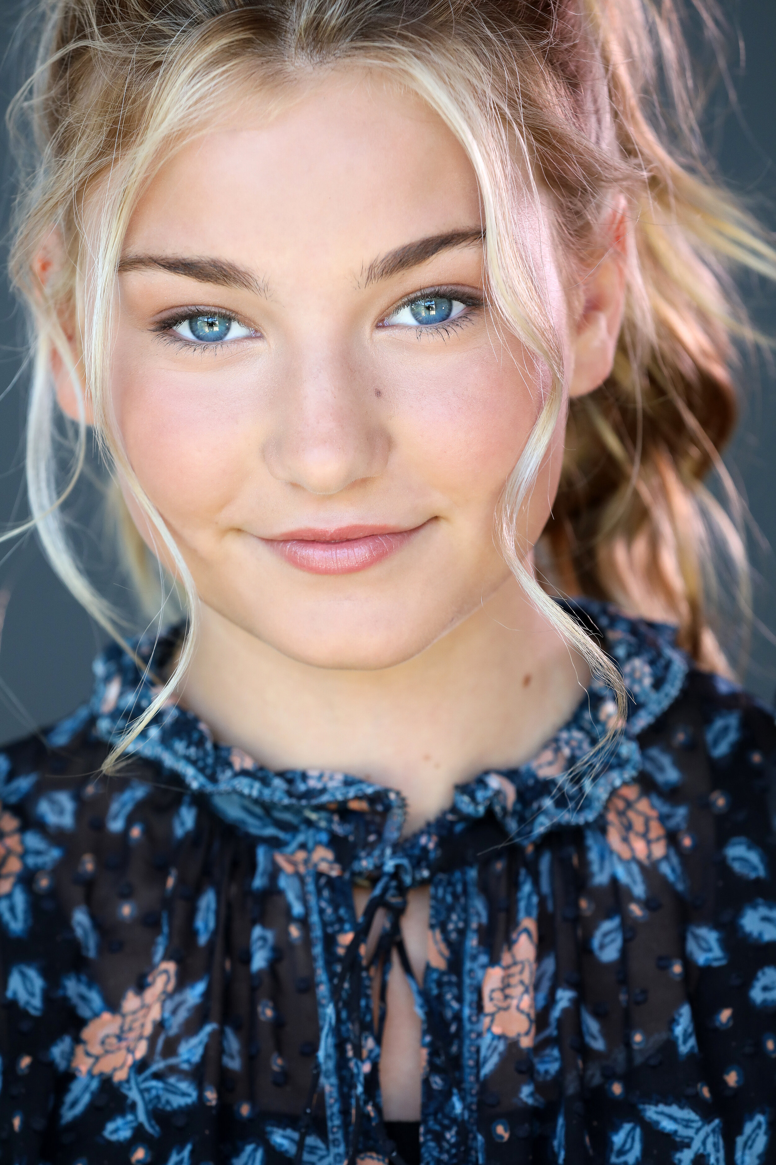 A young woman with blonde hair, blue eyes, and light makeup, looking at the camera with a slight smile, wearing a black floral top with blue and peach-colored flowers.