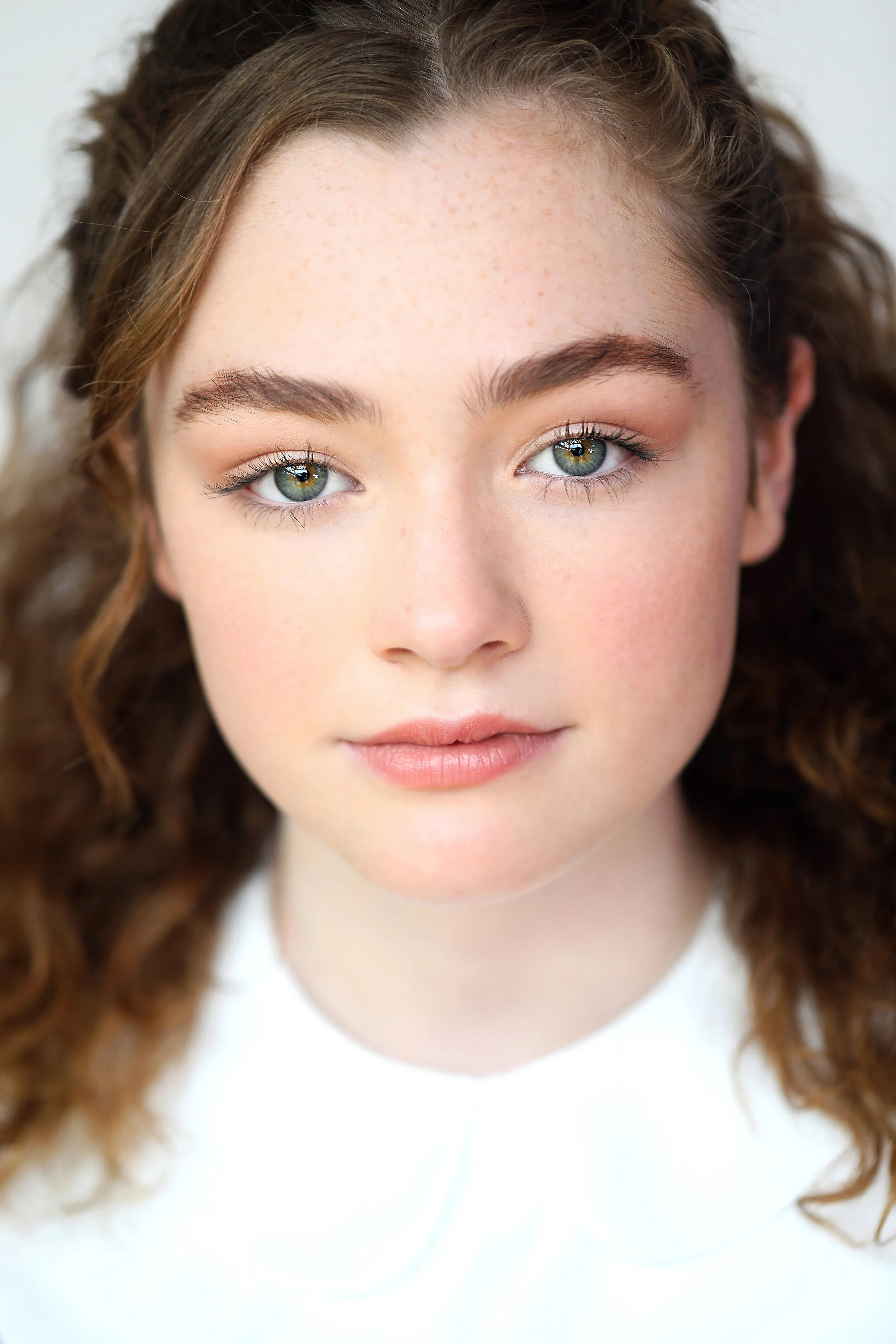 Close-up portrait of a young woman with curly brown hair, fair skin, and blue eyes, wearing a white top.