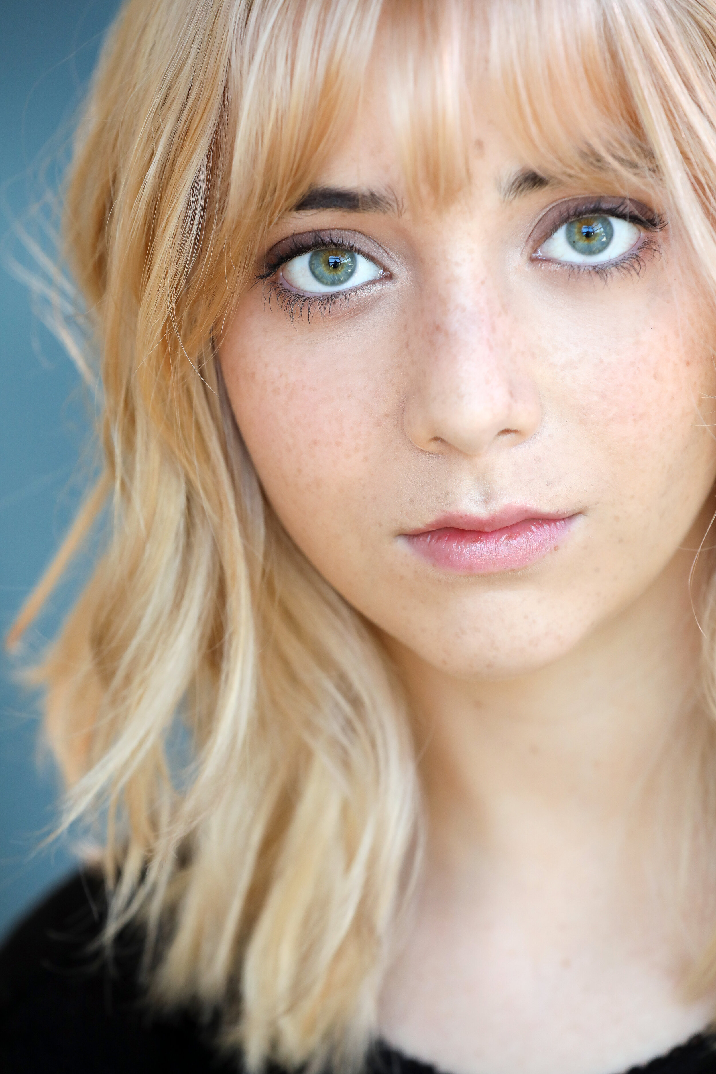 A close-up portrait of a young woman with blonde hair, green eyes, and freckles, looking directly at the camera.