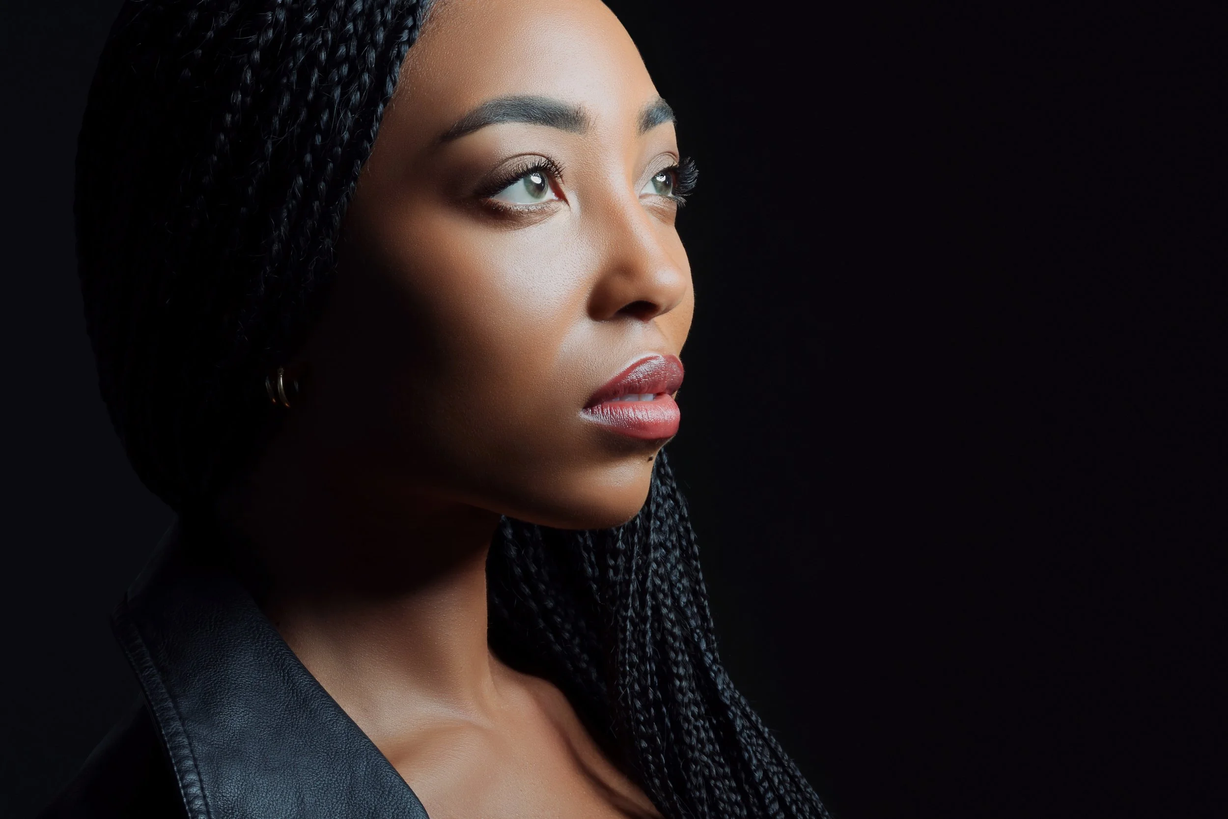 Close-up of a young woman with dark braided hair, wearing makeup, and looking slightly upward against a black background.